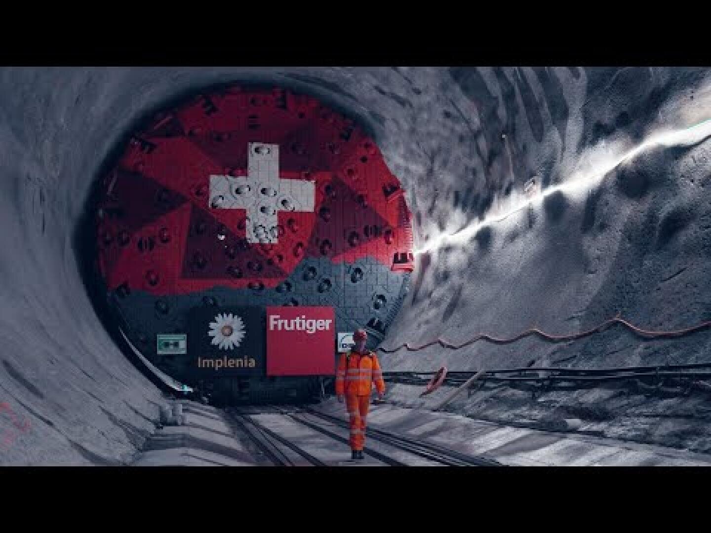 Eine Person in orangefarbener Schutzkleidung steht in einem Tunnel vor einer großen runden Maschine, die mit der Schweizer Flagge und den Firmenlogos von Implenia und Frutiger bemalt ist. Die Tunnelwände sind grau mit sichtbaren Kabeln.