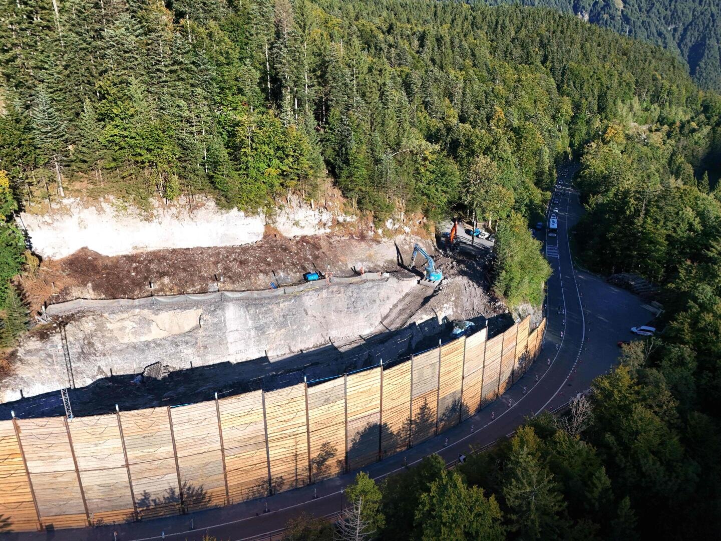 Luftaufnahme einer Baustelle an einer kurvenreichen Bergstraße, mit Baggern, die an einer hohen Stützmauer arbeiten, und dichtem Wald in der Umgebung.