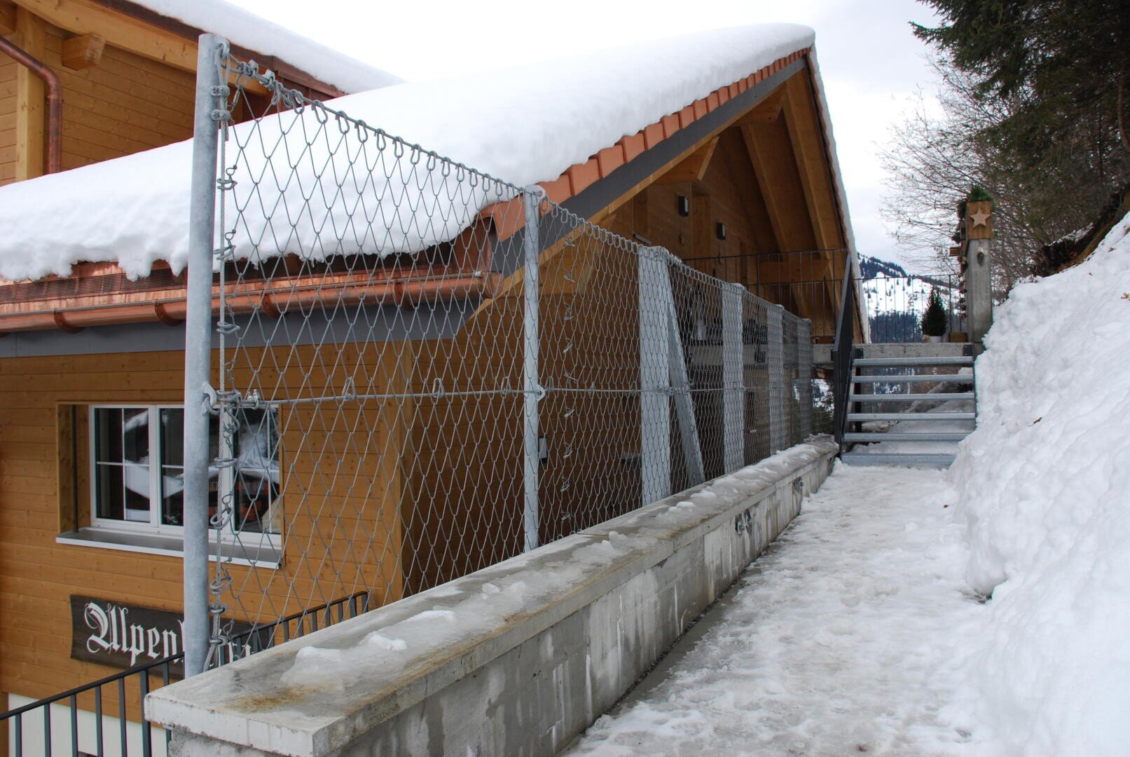 Ein schneebedeckter Weg mit einem Metallzaun führt an einem Holzgebäude mit Schrägdach vorbei. Am Ende des Weges führt eine Treppe nach oben, umgeben von Schnee und Bäumen.