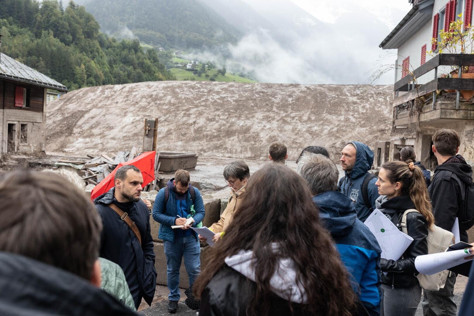 Eine Gruppe von Menschen steht in einem Bergdorf in der Nähe von Häusern und beobachtet und bespricht einen großen Erdrutsch, der den Hintergrund blockiert. Einige halten Papiere und Stifte in der Hand, während sie die Szene begutachten. Neblige Berge und bewölkter Himmel sind zu sehen.