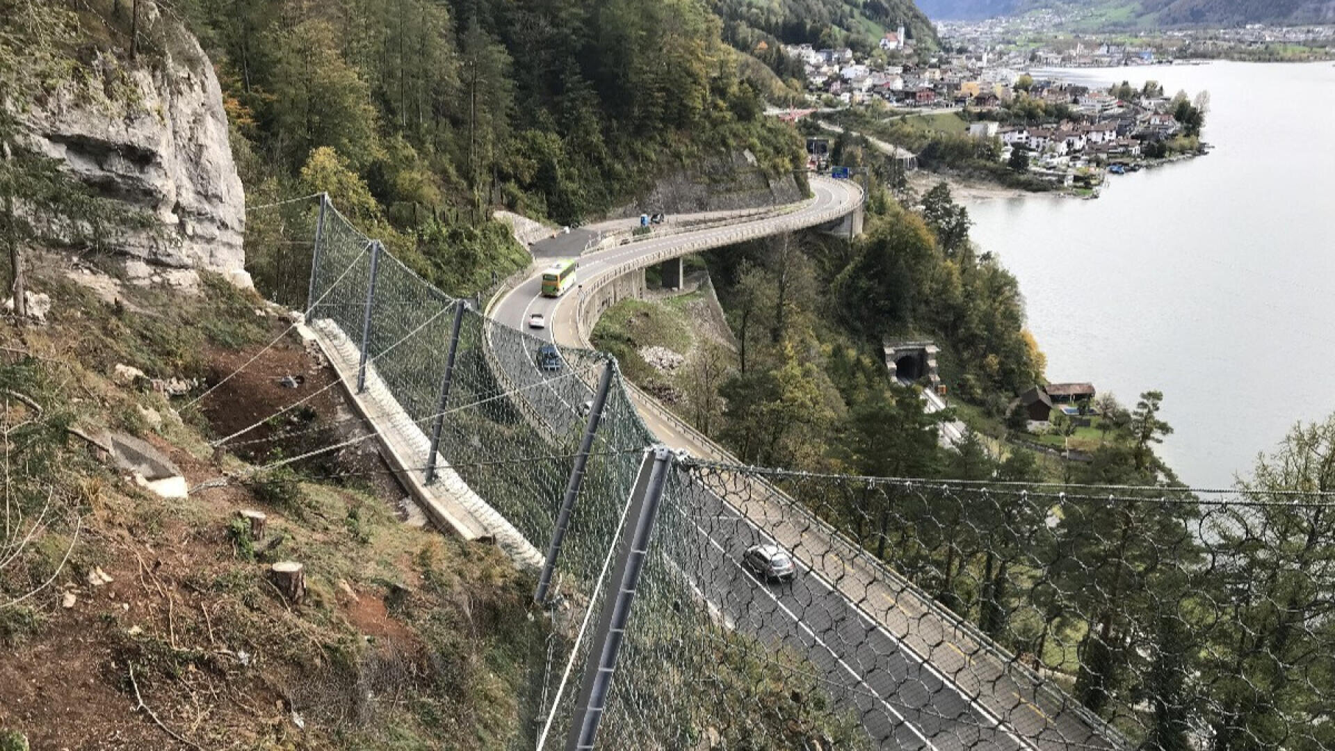 An einem steilen Abhang neben einer kurvenreichen Straße in der Nähe eines Sees ist eine Steinschlagbarriere aus Metall installiert, unter der Autos fahren und in der Ferne ein Dorf zu sehen ist. Das Gebiet ist von Bäumen und felsigen Klippen umgeben.