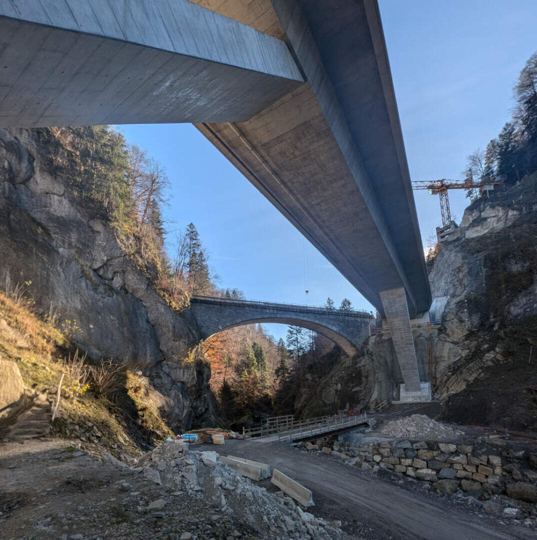 Zwei Brücken überqueren ein felsiges Tal: oben eine moderne Betonbrücke und unten eine ältere Steinbogenbrücke. Entlang der Klippen wachsen Bäume, und unten sind ein Baukran und Fahrzeuge auf der Baustelle zu sehen.