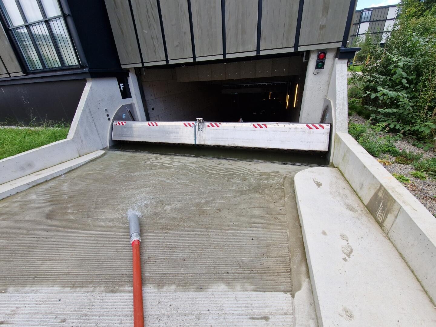 Eine Schranke versperrt die Einfahrt zu einer Tiefgarage, während auf der Rampe Wasser gespritzt wird oder fließt. Ein rot-weiß gestreiftes Tor und eine Ampel sind zu sehen, auf der rechten Seite befindet sich eine Grünanlage.
