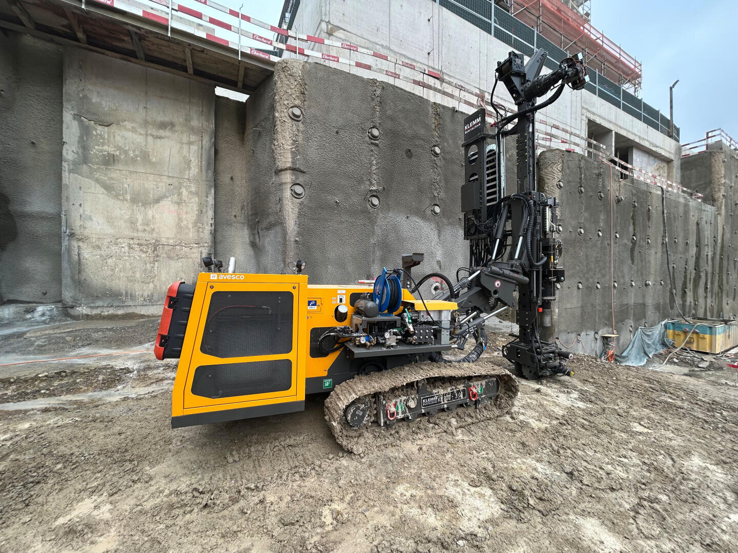 Eine gelbe Bohrmaschine mit Raupenfahrwerk steht auf einer schlammigen Baustelle in der Nähe hoher, unfertiger Betonwände. Im Hintergrund sind Gerüste und rote Sicherheitsbarrieren zu sehen.