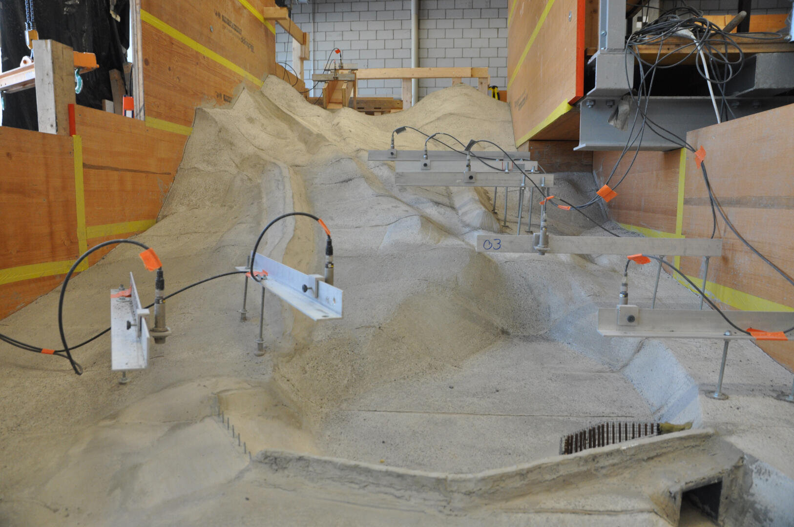 Ein Laboraufbau mit Sensoren, die auf Metallbügeln über einem Betonmodell eines unebenen Geländes montiert sind, wahrscheinlich für Tests oder zur Simulation von Umweltbedingungen. An den Seiten sind hölzerne Barrieren und Verkabelung sichtbar.
