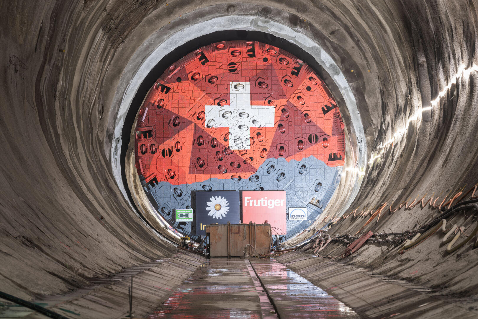 Eine große Tunnelbohrmaschine mit einem Schneidkopf in Form der Schweizer Flagge steht am Ende eines im Bau befindlichen Tunnels. Die Wände und der Boden des Tunnels sind mit Baustellenlampen und -geräten ausgekleidet.