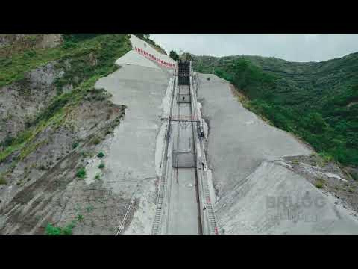 Ein langes, steiles Betonbauwerk fällt zwischen grünen Hügeln ab und ähnelt einer großen Entwässerungsrutsche oder einer Erdrutschbarriere, mit Geländern und Wegen in der Mitte.