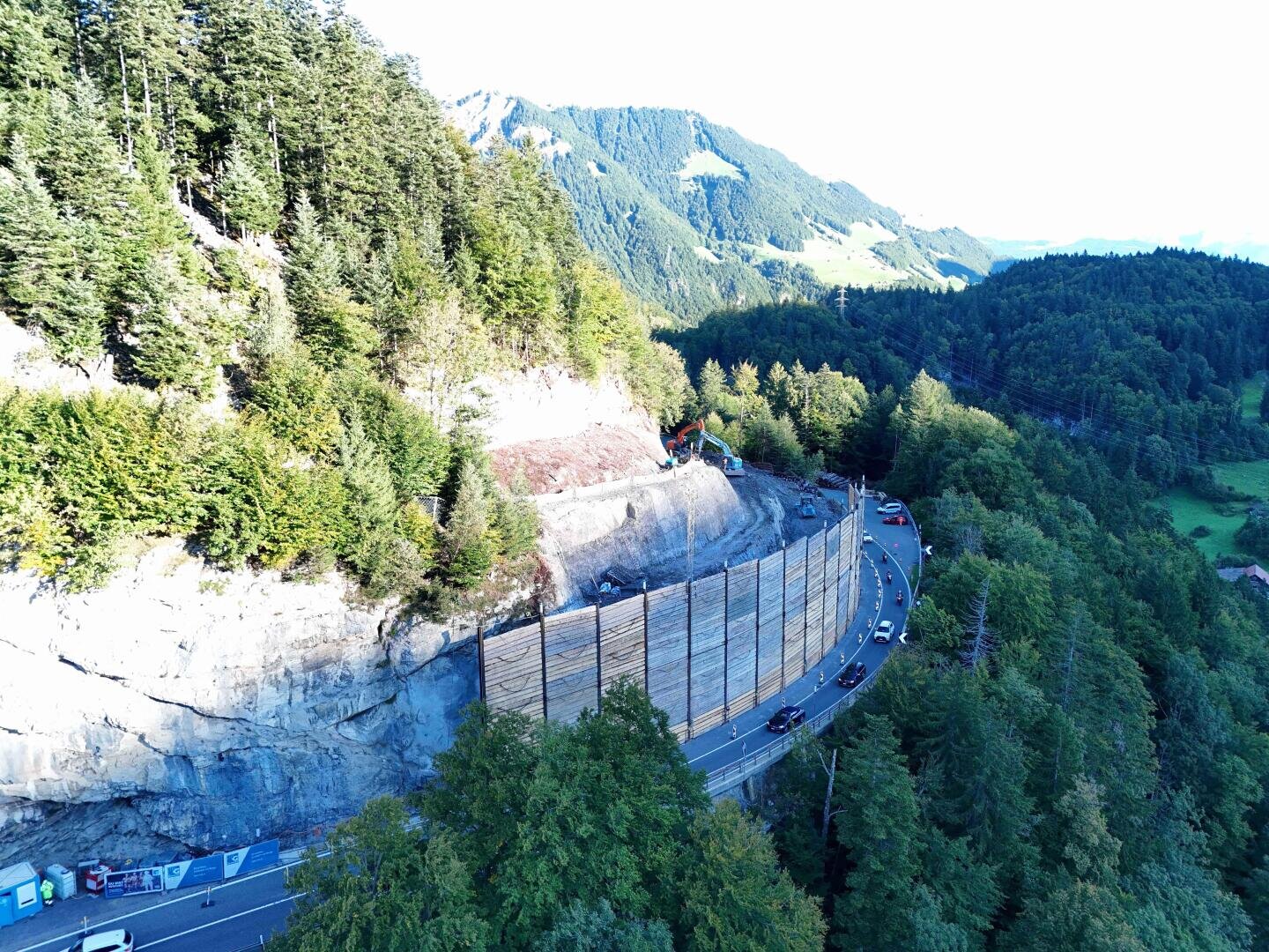 Eine kurvenreiche Bergstraße schlängelt sich durch einen bewaldeten Berghang, über dem eine Baustelle den Hang befestigt. Mehrere Autos fahren auf der Straße, die von hohen Schutzzäunen begrenzt wird. Grüne Bäume und Berge umgeben das Gebiet.