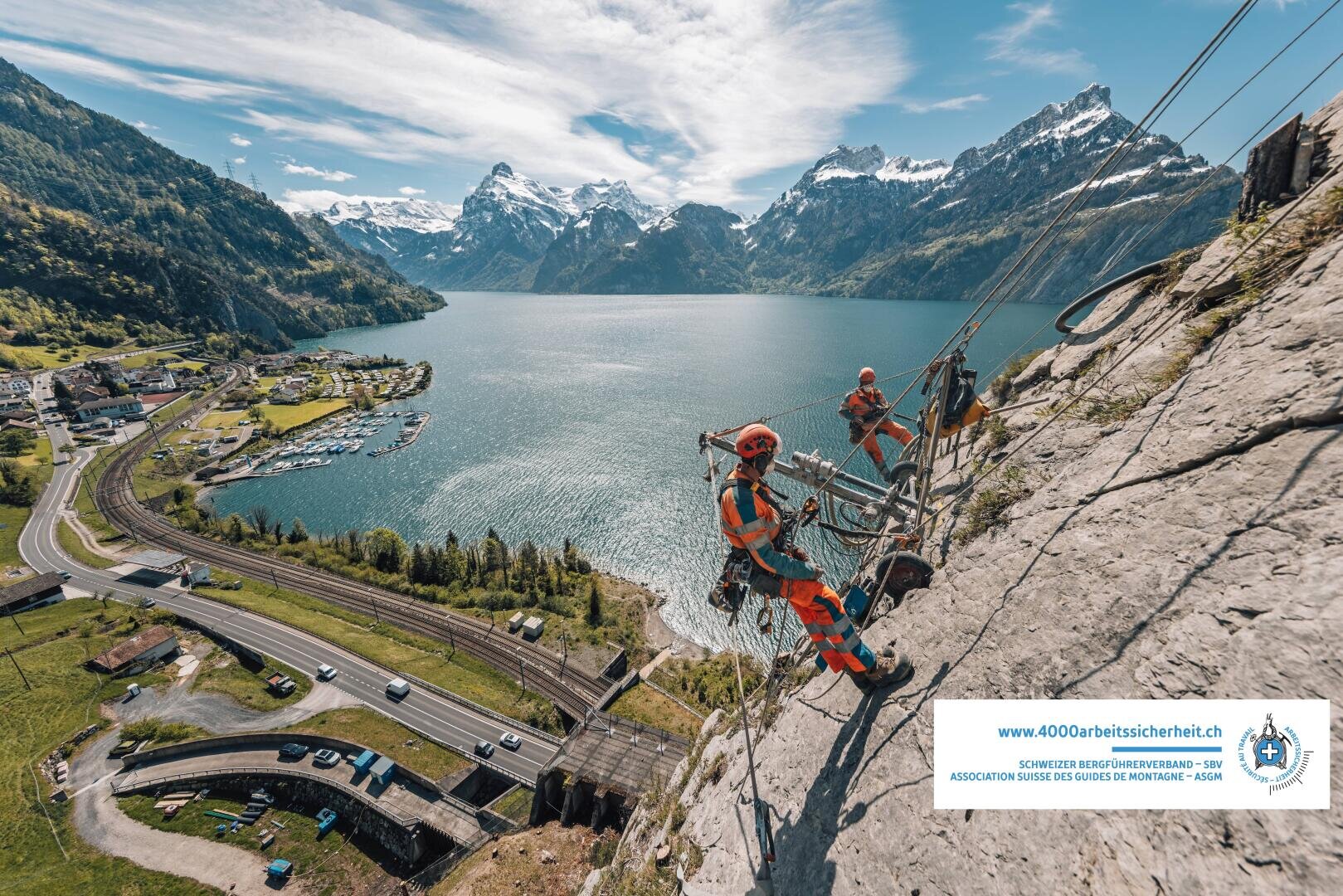 Zwei Arbeiter in orangefarbenen Schutzanzügen und Helmen führen Wartungsarbeiten an einem steilen Berghang oberhalb eines malerischen Sees durch, während im Hintergrund eine Stadt, Straßen und schneebedeckte Berge zu sehen sind.