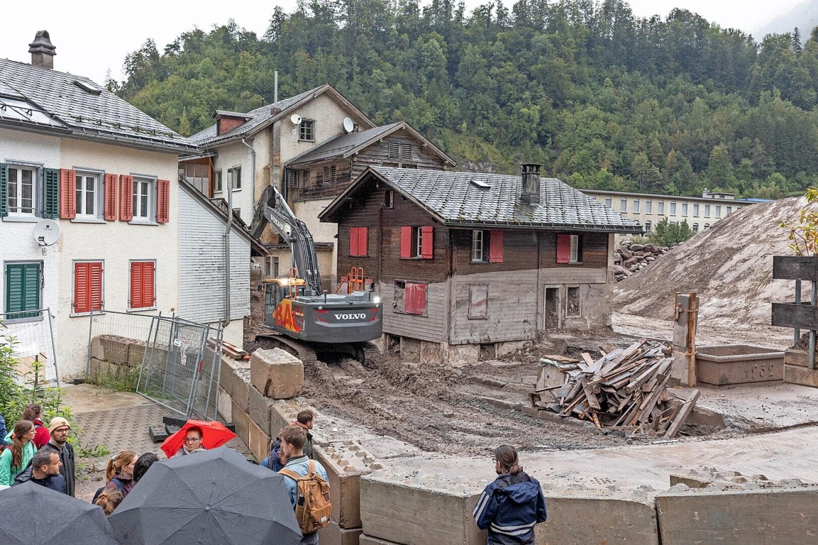 Eine Gruppe von Menschen sieht zu, wie ein Volvo-Bagger Schlamm und Schutt um die beschädigten Häuser in einem Dorf nach einem Erdrutsch beseitigt, mit schlammigem Boden, Bauabsperrungen und einer Kulisse aus grünen, bewaldeten Hügeln.