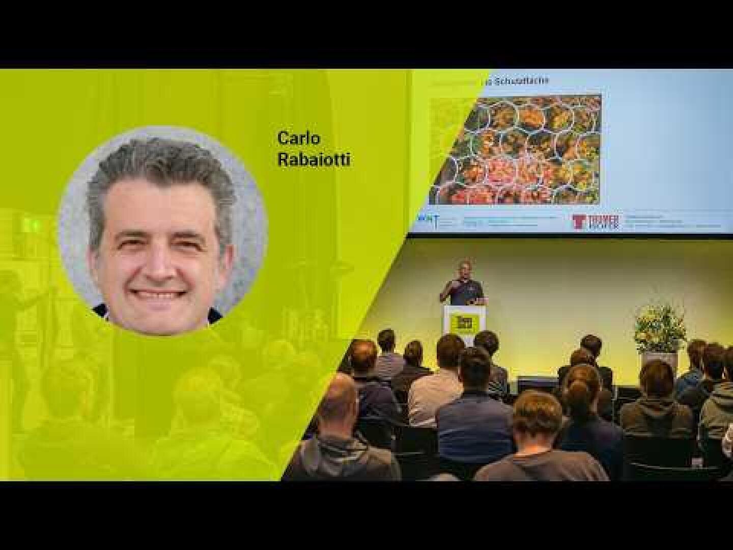 Eine Präsentationsveranstaltung mit dem Foto und dem Namen von Carlo Rabaiotti auf einer gelben Grafik; er steht auf einem Podium und spricht zu einem sitzenden Publikum, während hinter ihm ein großes Bild von Lebensmitteln auf dem Bildschirm erscheint.