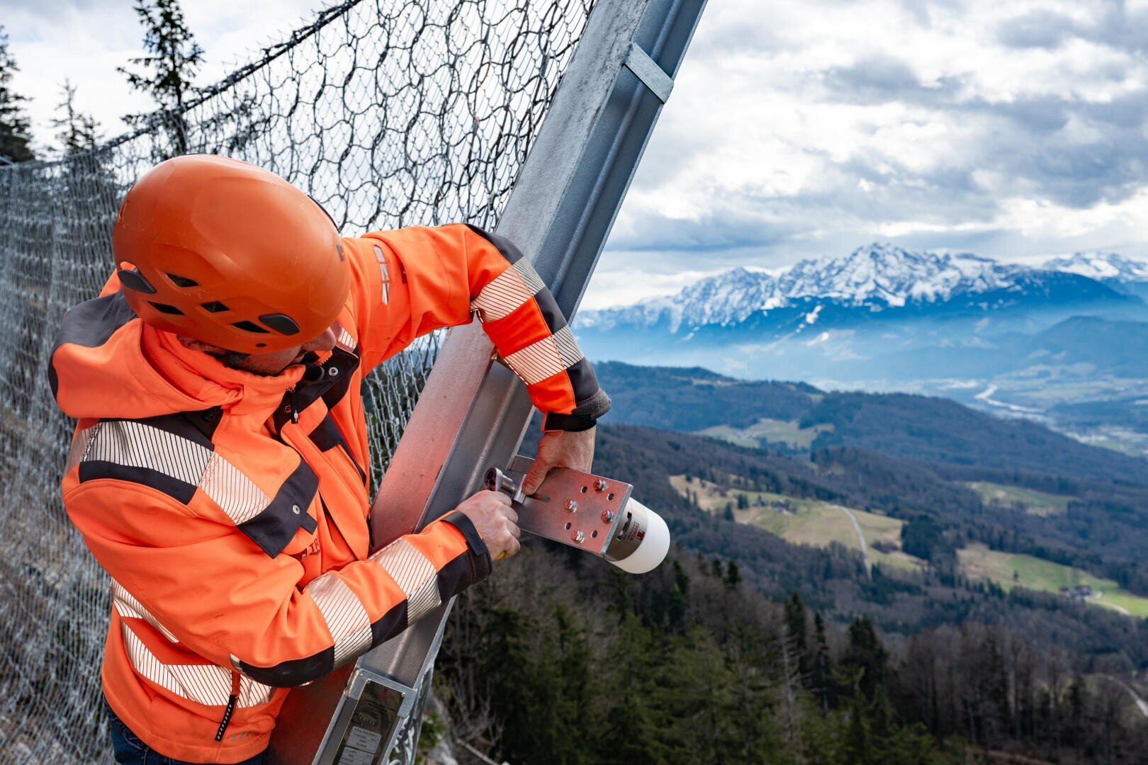 Ein Arbeiter in orangefarbener Sicherheitsjacke und mit Helm befestigt einen Metallpfosten an einem Drahtzaun an einem steilen Abhang, während im Hintergrund Berge und ein bewaldetes Tal zu sehen sind.