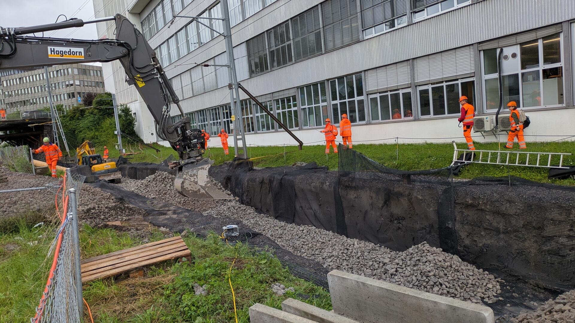 Bauarbeiter in orangefarbener Schutzkleidung arbeiten in der Nähe eines Gebäudes mit einem großen Bagger, der Steine und Netze in einem langen Graben verlegt, während Geräte und Absperrungen die Baustelle umgeben.