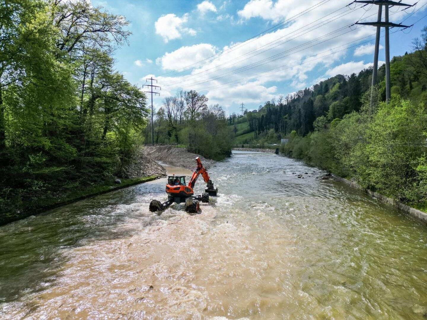 Ein orangefarbener Amphibienbagger arbeitet in einem seichten Fluss, der von grünen Bäumen und Hügeln umgeben ist, unter einem blauen Himmel mit Wolken und Stromleitungen über dem Kopf.