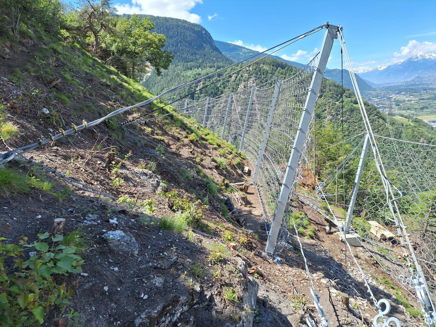 Ein Steinschlagschutzzaun aus Metall ist an einem steilen, felsigen Berghang mit grüner Vegetation installiert. Im Hintergrund sind Berge und ein Tal unter einem blauen Himmel mit vereinzelten Wolken zu sehen.