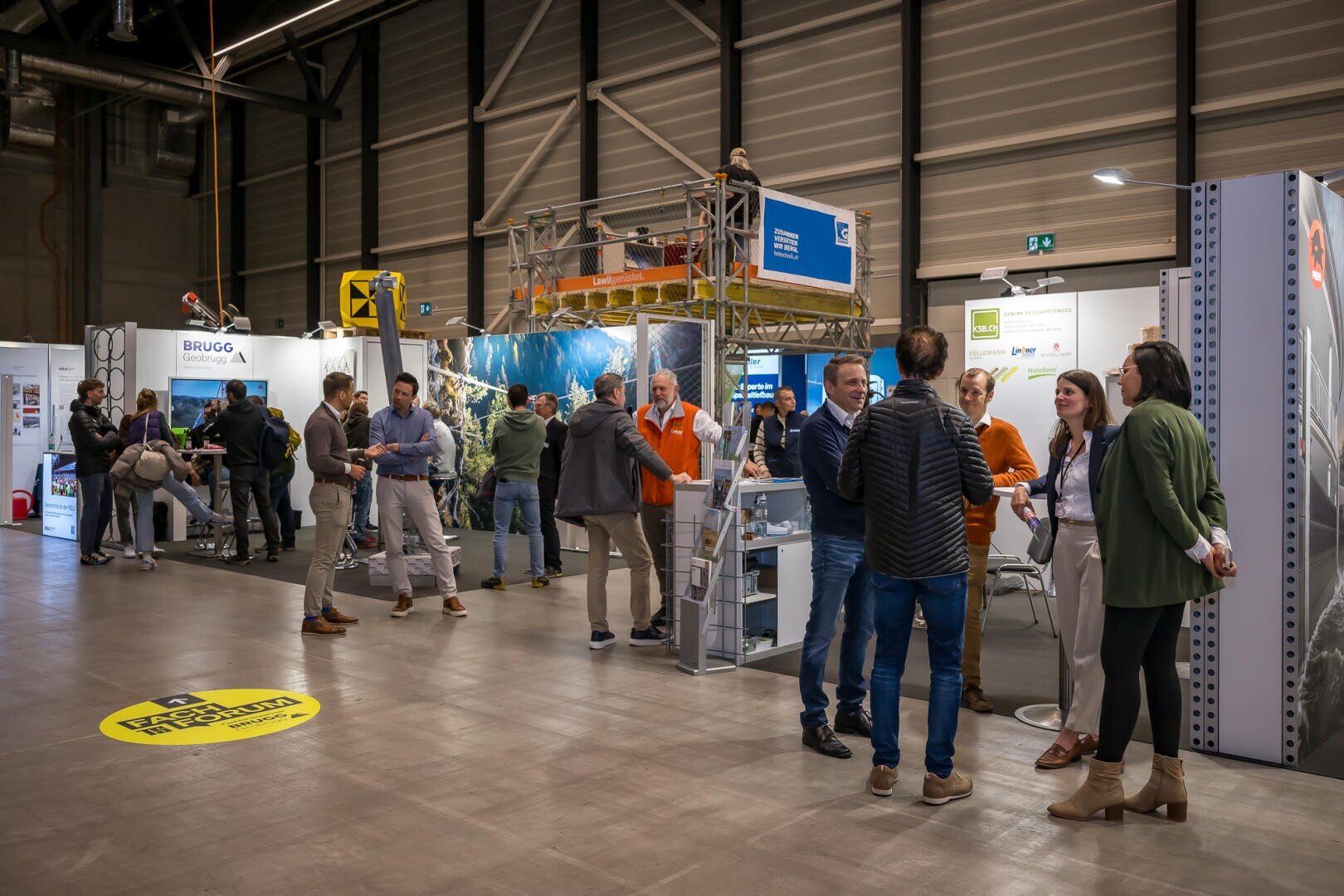 Auf einer Messe mit Messeständen und Auslagen sind die Menschen in Kontakt. Einige stehen in Gruppen und unterhalten sich, während andere die Stände mit Industrieausrüstungen und Informationsmaterial in einer geräumigen, gut beleuchteten Halle erkunden.