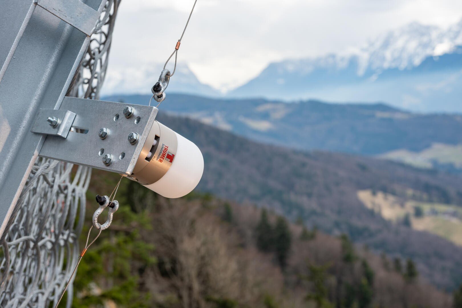 Nahaufnahme eines Sensors oder Überwachungsgeräts an einem Metallzaun mit Blick auf eine Berglandschaft mit Wäldern und schneebedeckten Gipfeln im Hintergrund.