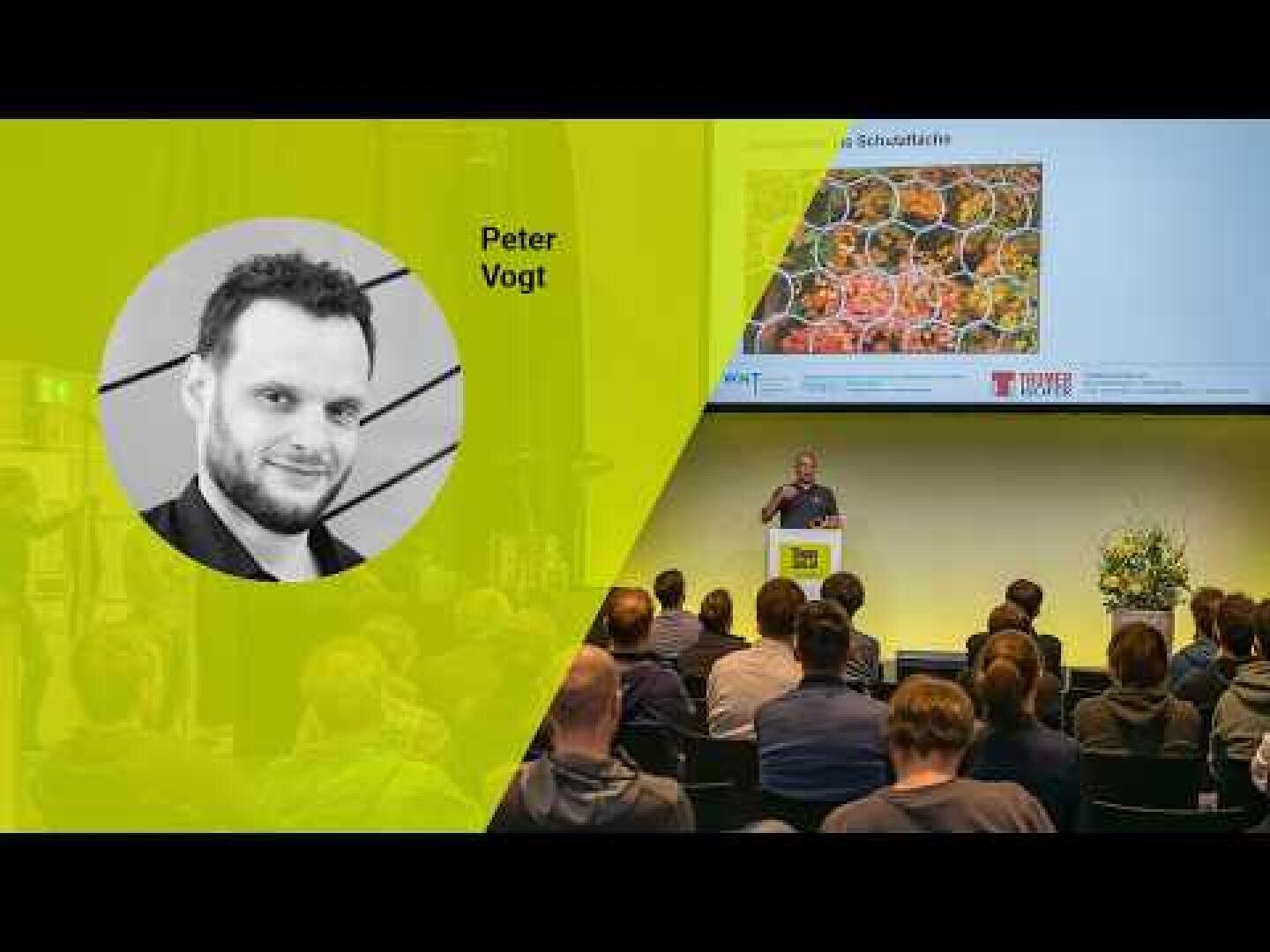 Es findet eine Präsentation vor einem Publikum statt. Auf der Leinwand sind ein großes Foto eines Mannes mit der Aufschrift Peter Vogt und Dias mit bunten Bildern zu sehen. Das Publikum sitzt und konzentriert sich auf den Redner.
