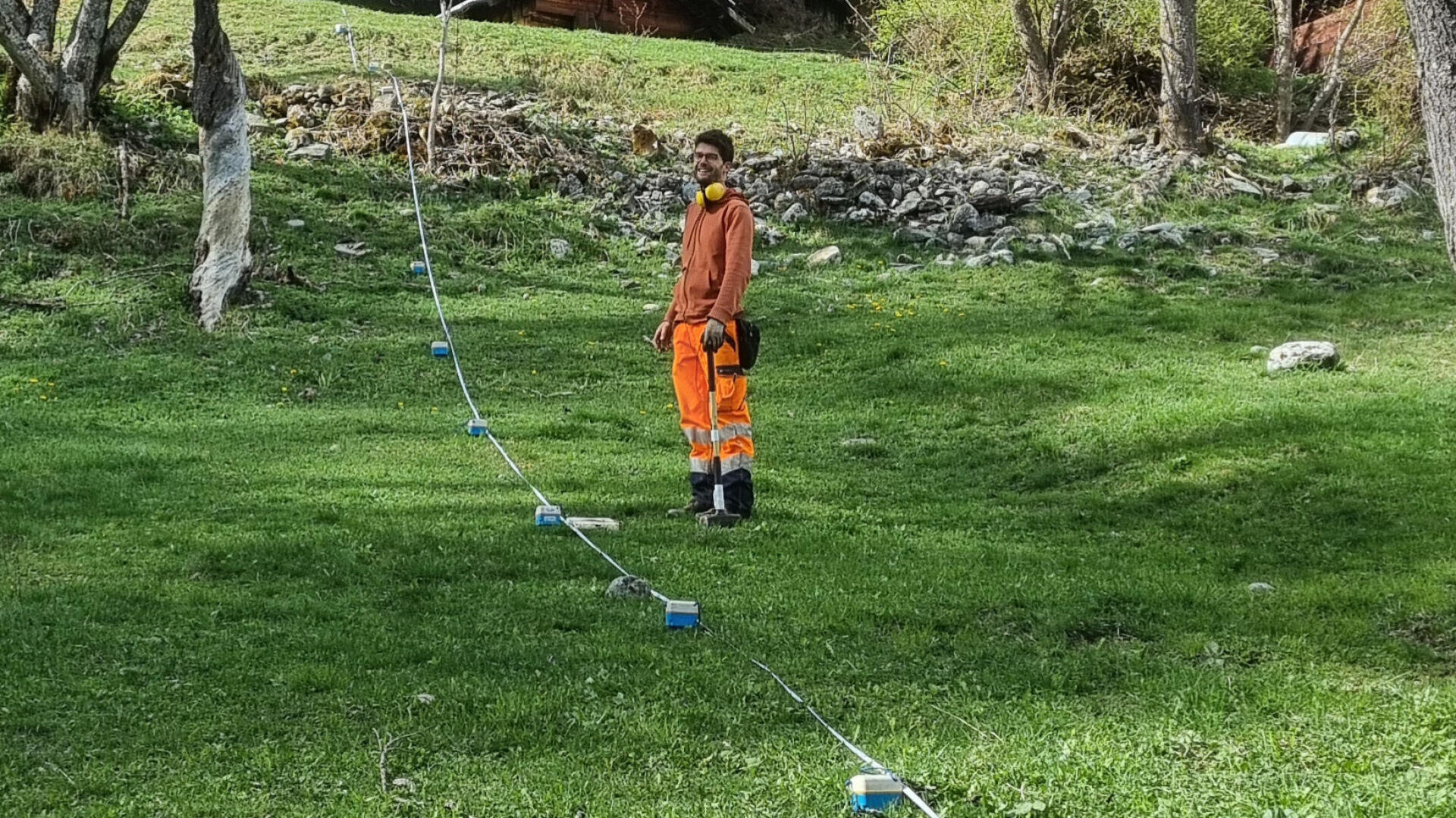 Eine Person mit orangefarbener Schutzkleidung und Gesichtsmaske steht auf einem grasbewachsenen Gelände neben einer Reihe blauer und weißer Sensorgeräte, die in Abständen auf dem Boden verteilt sind.