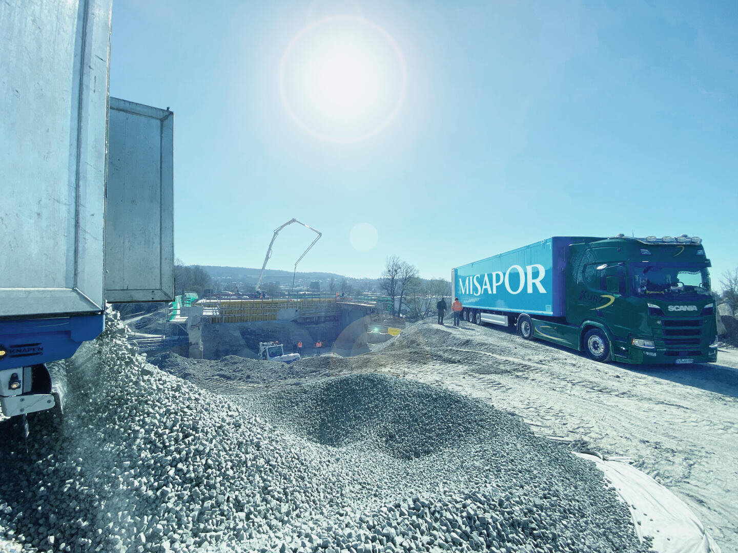 Ein heller, sonniger Tag auf einer Baustelle mit zwei großen Lastwagen - ein blauer und ein grüner mit der Aufschrift MISAPOR. Schotterhaufen und Maschinen sind zu sehen, im Hintergrund Arbeiter und über ihnen der blaue Himmel.