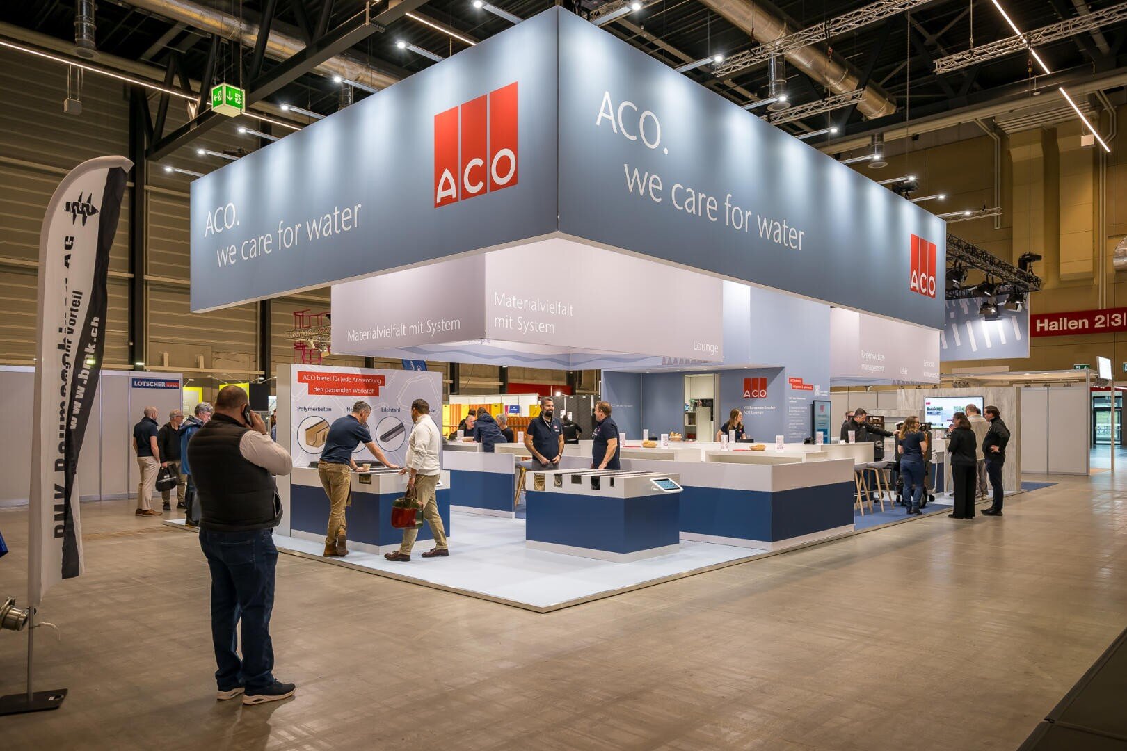 Auf einem modernen Messestand von ACO mit dem Slogan "We care for water" werden Produkte für die Wasserwirtschaft ausgestellt. Mehrere Personen unterhalten sich mit dem Personal und erkunden den Stand in einer hell erleuchteten Kongresshalle.