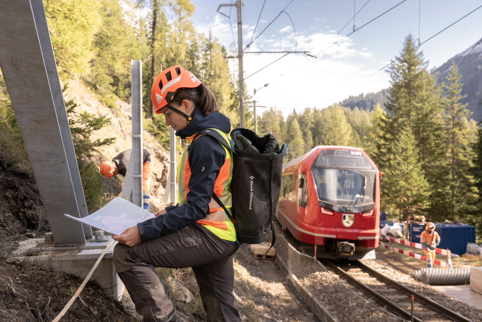 Ein Bauarbeiter in Schutzkleidung prüft eine Karte neben einem Bahngleis in einer bewaldeten, gebirgigen Gegend, während sich im Hintergrund ein roter Zug nähert. Andere Arbeiter und Baumaterialien sind in der Nähe.