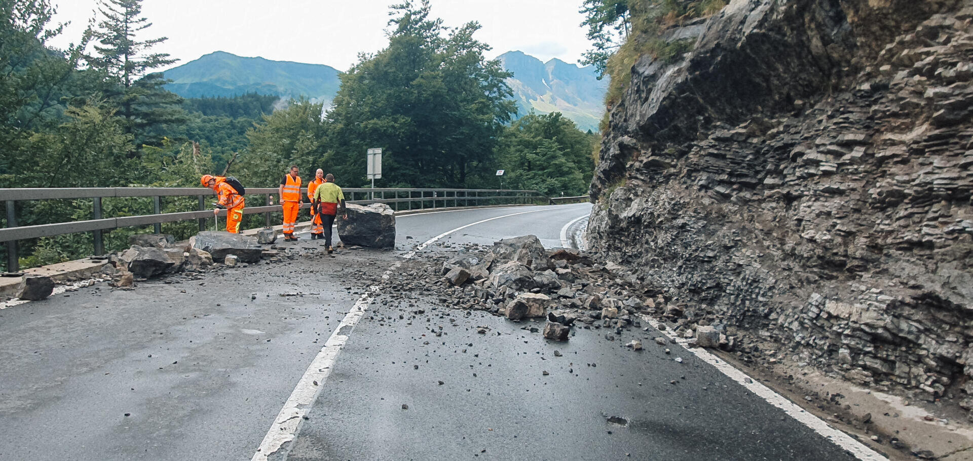 Straßenarbeiter in hellen Uniformen befreien eine Bergstraße nach einem Erdrutsch von herabgefallenen Steinen und Geröll, mit einer Leitplanke und bewaldeten Hügeln im Hintergrund.