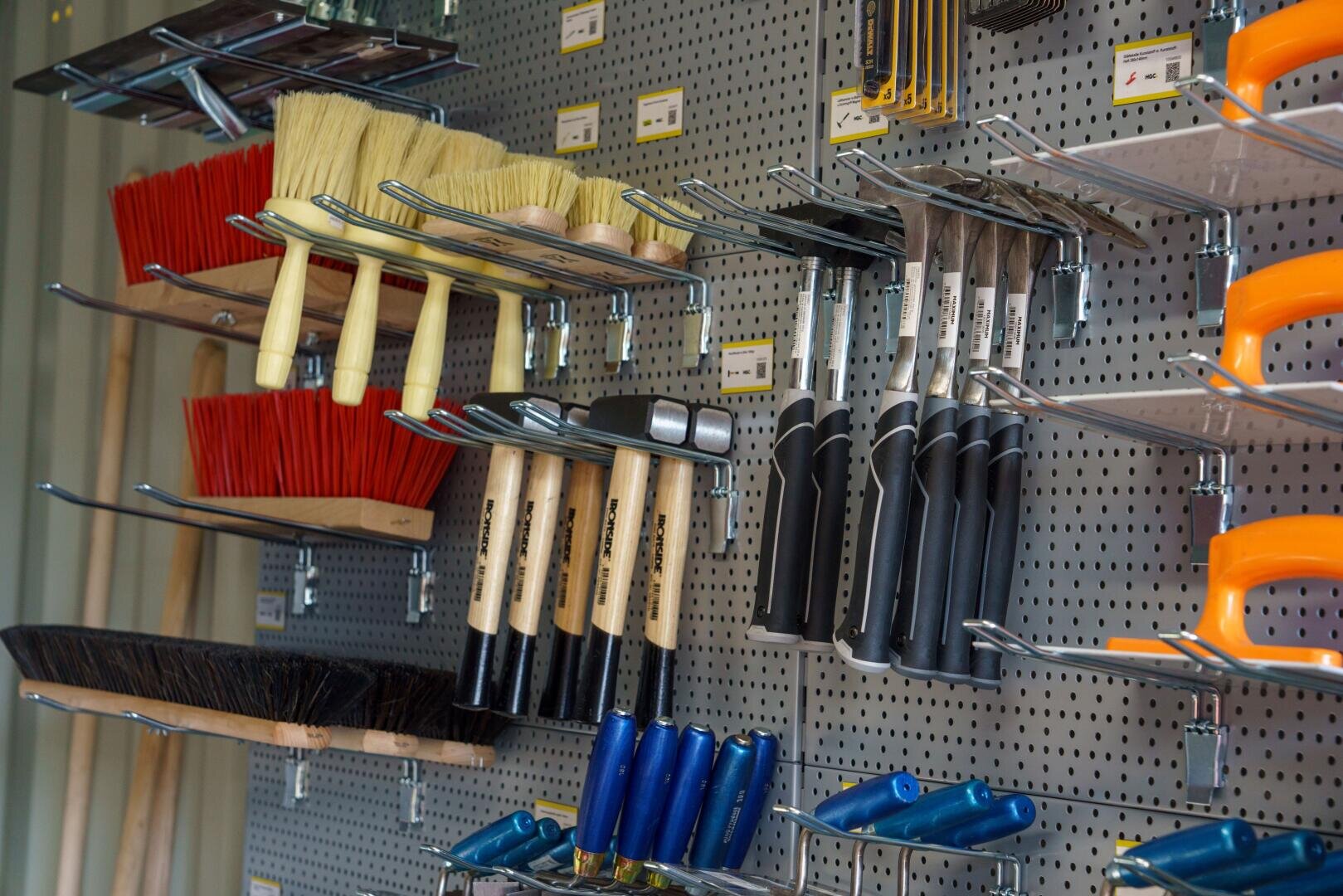 Verschiedene Handwerkzeuge, darunter Bürsten, Hämmer, Sägen und Pinsel, sind ordentlich an Metallhaken aufgehängt, die an einer Lochplattenwand in einem Baumarkt befestigt sind.