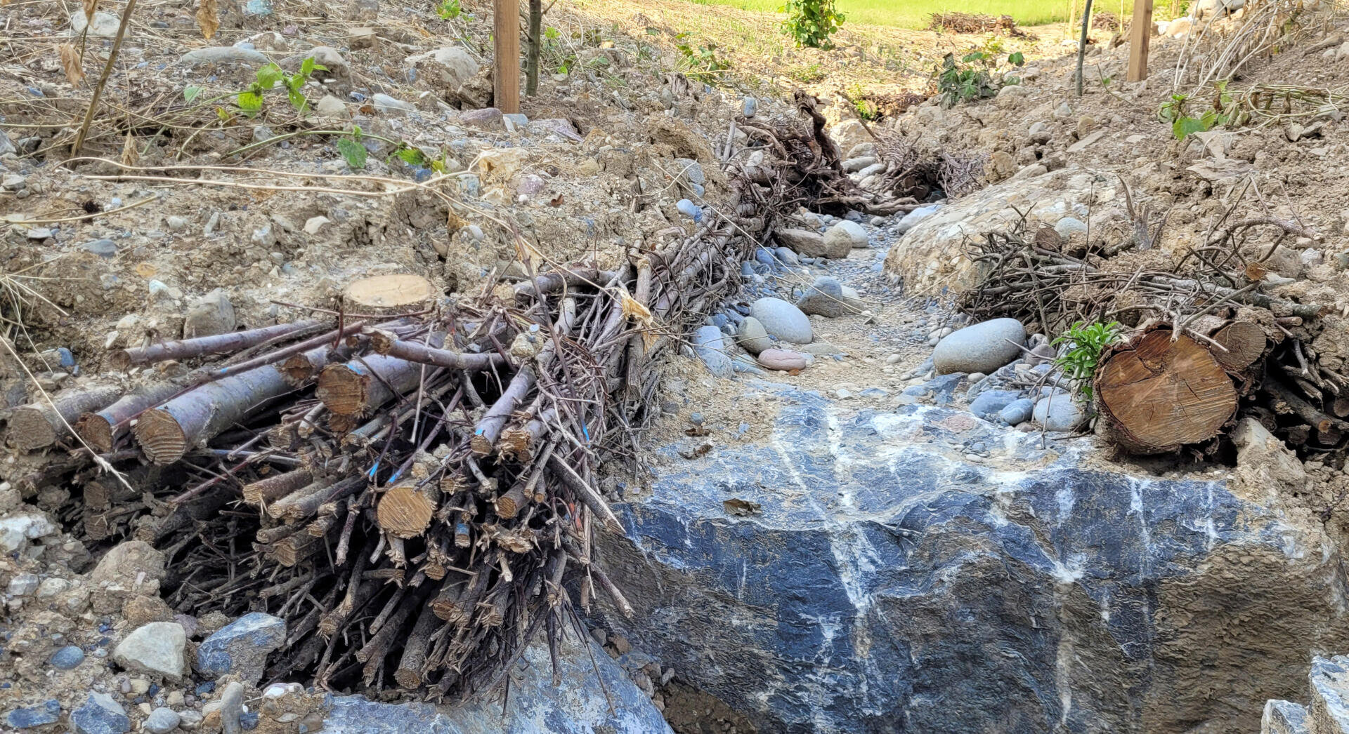 Ein kleiner Graben im felsigen Boden ist auf beiden Seiten mit gebündelten Stöcken und Ästen verstärkt, und am Boden liegen einige glatte Steine. Zwischen der Erde und den Steinen wachsen spärliche Grünpflanzen.