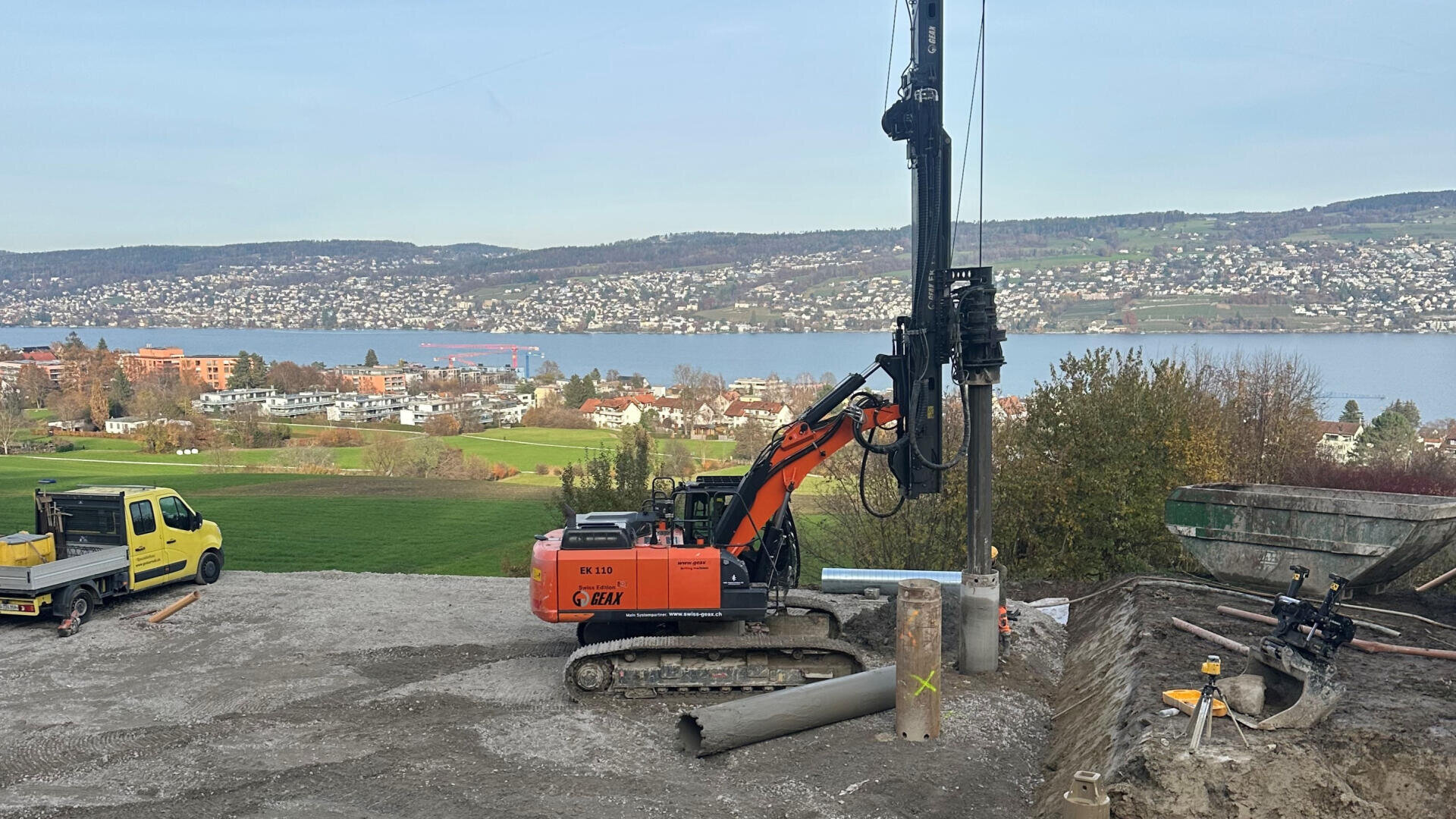 Ein orangefarbener Bagger bohrt auf einer Baustelle mit Blick auf einen großen See in den Boden, während in der Nähe ein gelber Lieferwagen parkt und im Hintergrund eine Stadt und Hügel zu sehen sind.