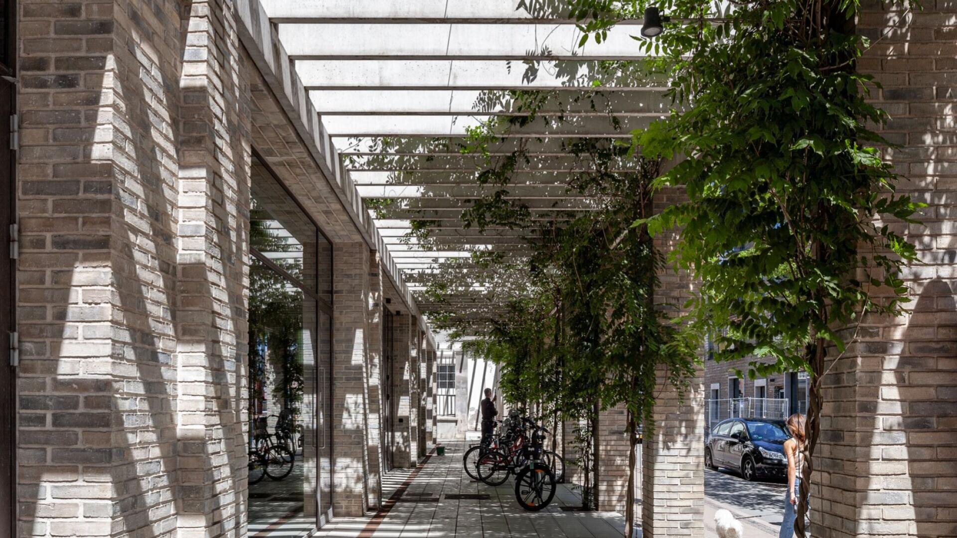 Ein moderner Gehweg mit Backsteinsäulen und einer weißen Pergola über dem Kopf, gesäumt von grünen Kletterpflanzen. An der Seite sind Fahrräder geparkt, und rechts geht eine Person spazieren. Das Sonnenlicht wirft Schatten, und draußen sind Autos zu sehen.