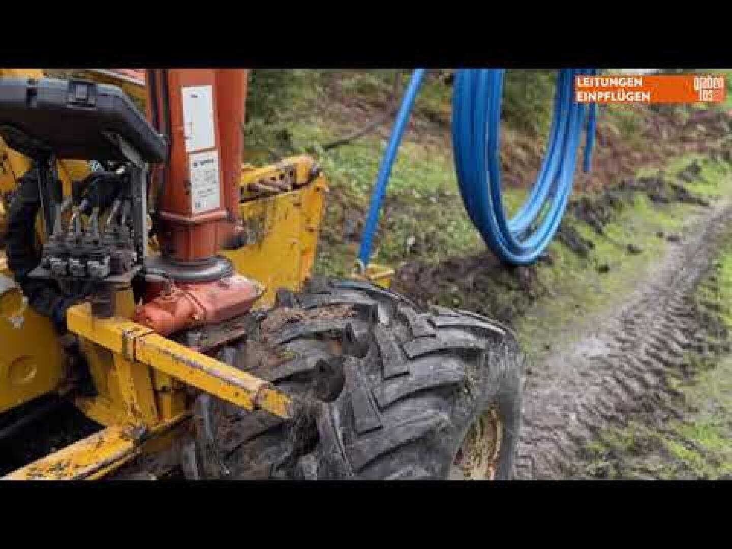 Nahaufnahme eines gelben Baufahrzeugs mit einem schlammigen Reifen auf einem Feldweg. In der Nähe der Maschine sind blaue Rohre aufgerollt, und der Boden weist Reifenspuren auf. Im Hintergrund sind Gras und Bäume zu sehen.