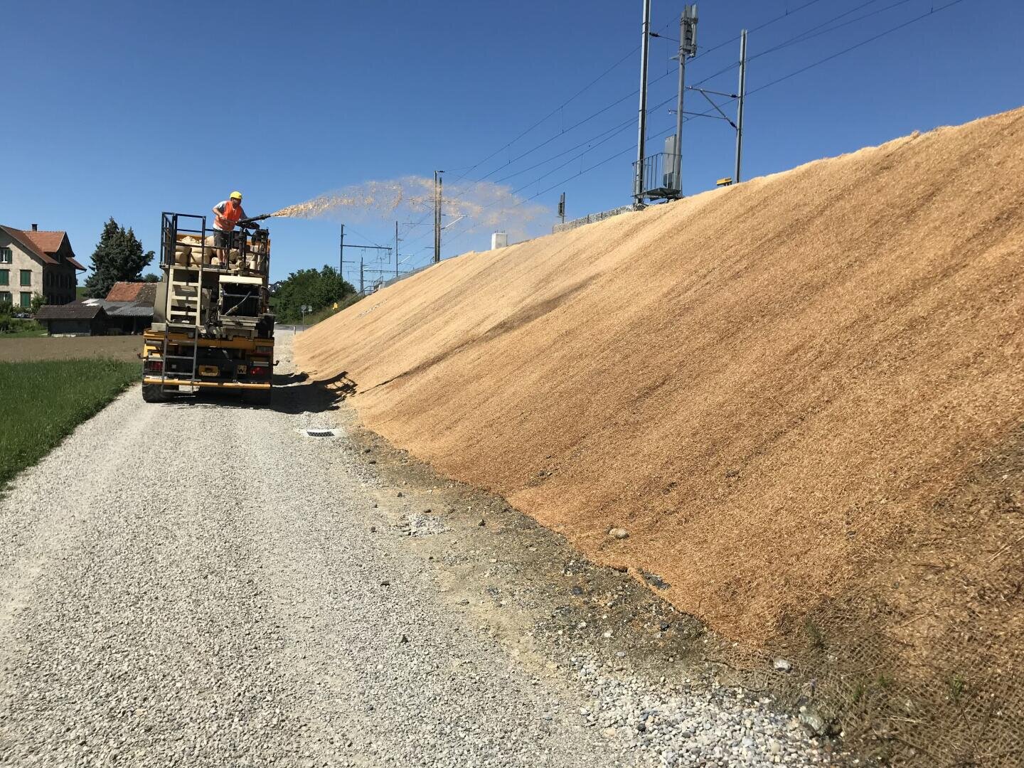 Ein Arbeiter auf einem Lastwagen sprüht ein hellbraunes Material auf eine steile Böschung neben einer Schotterstraße in der Nähe von Stromleitungen, im Hintergrund sind Häuser und Gras zu sehen, der Himmel ist blau.