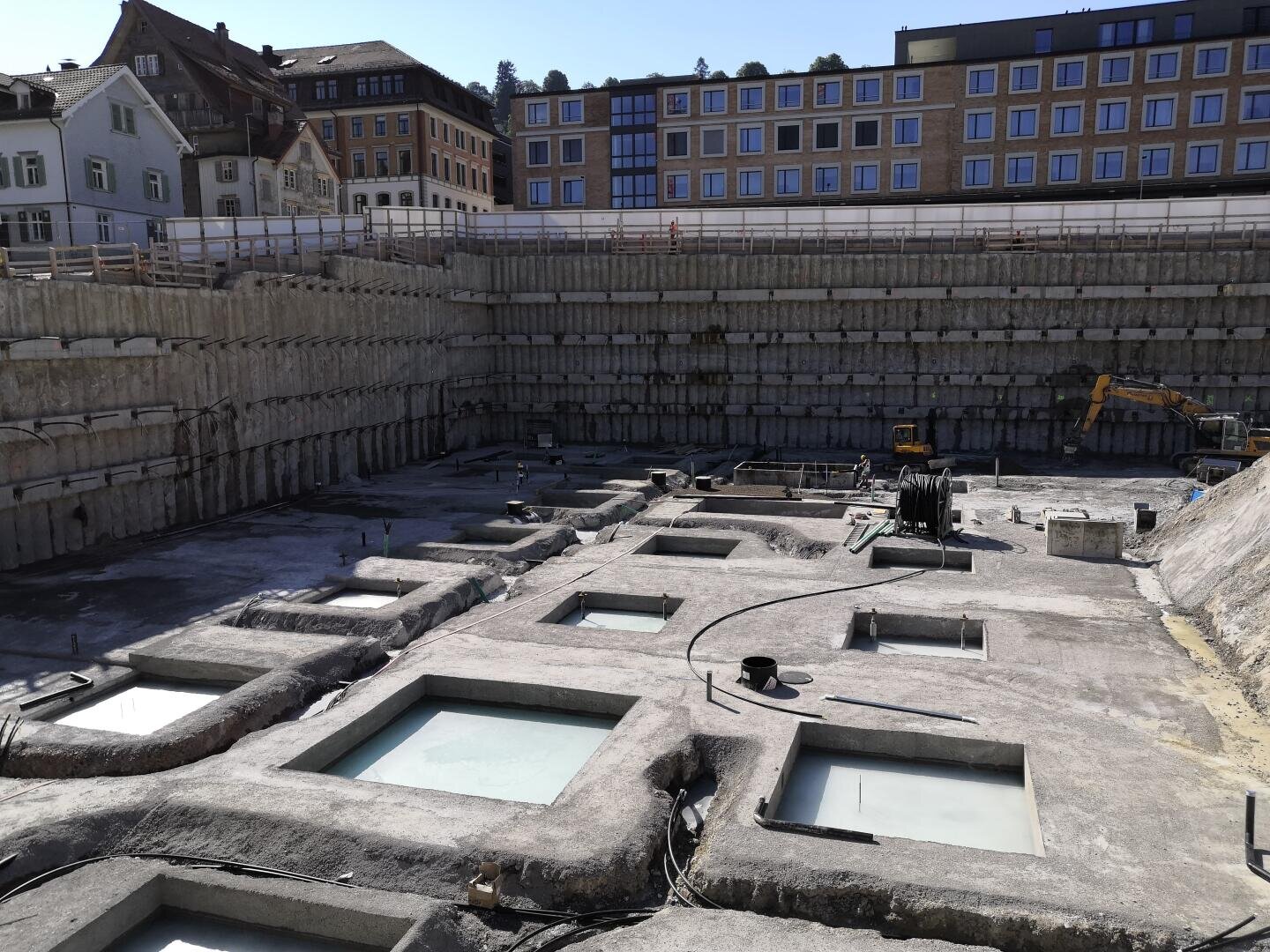Eine große Baustelle mit tiefen Baugruben und Stützkonstruktionen; im Hintergrund sind bei klarem Himmel moderne Gebäude und ältere Häuser zu sehen.