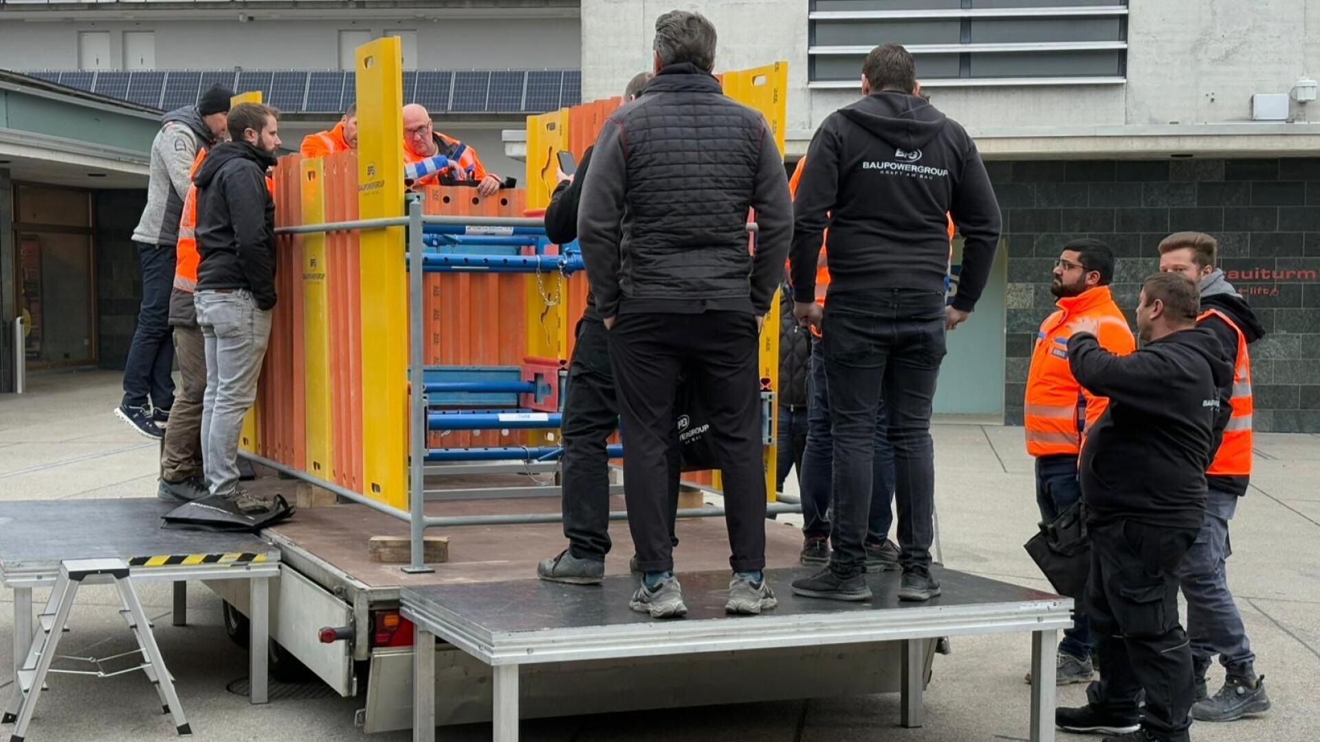 Eine Gruppe von Arbeitern, einige in orangefarbenen Sicherheitswesten, steht um die offene Ladefläche eines Lastwagens herum und hantiert mit bunten Metallstrukturen. Die Szene scheint sich im Freien in der Nähe eines modernen Gebäudes abzuspielen.