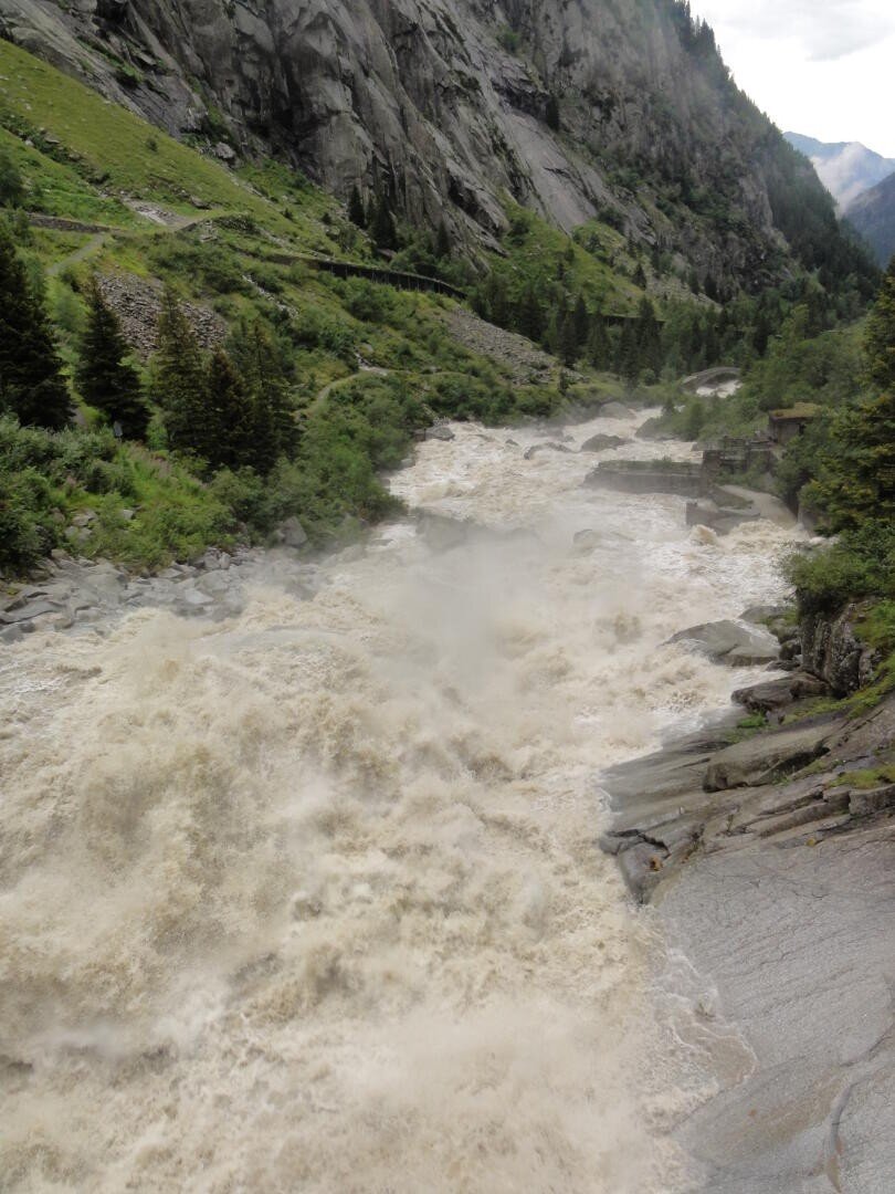 Ein mächtiger Fluss mit schäumenden Stromschnellen rauscht durch eine felsige, gebirgige Landschaft mit dichten grünen Bäumen und steilen Klippen unter einem bewölkten Himmel.