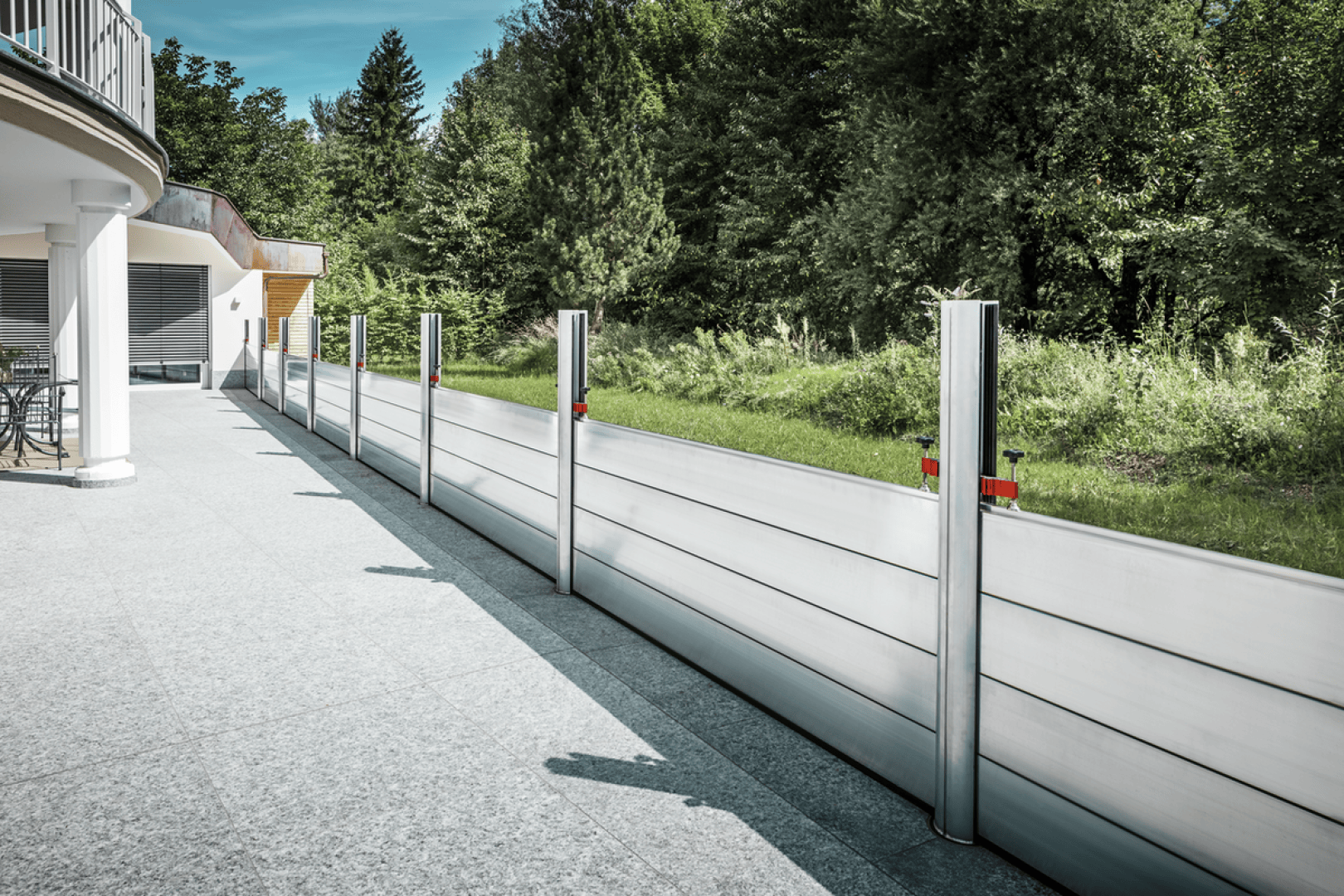 Eine Hochwasserschutzwand aus Aluminium, die am Rand des Innenhofs eines modernen Gebäudes installiert wurde, mit grünem Gras und dichten Bäumen im Hintergrund an einem klaren Tag.