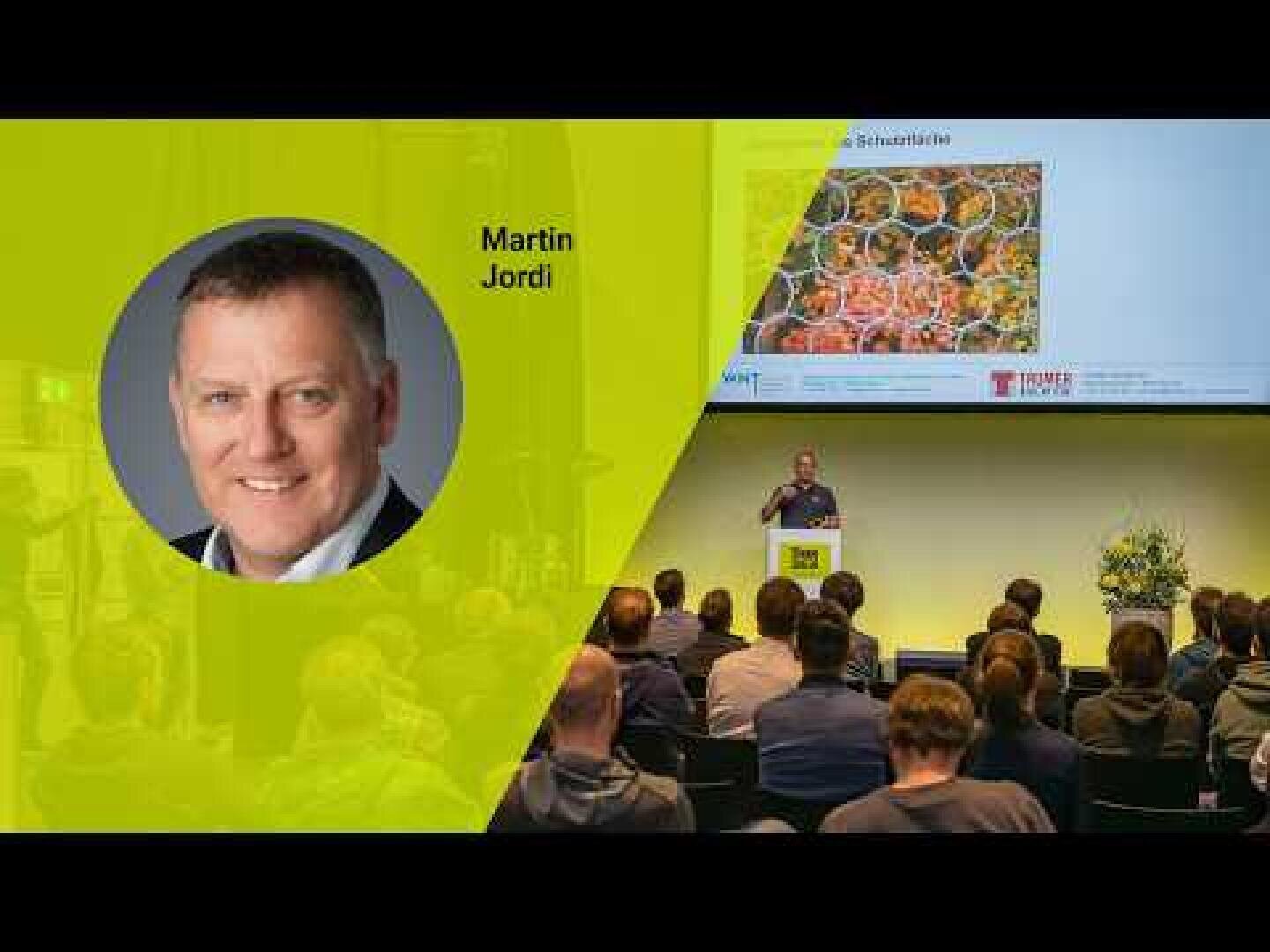 Ein Redner hält einen Vortrag vor Publikum in einem Konferenzraum. Auf der linken Seite zeigt ein großes gelb-grünes Overlay ein rundes Foto eines lächelnden Mannes mit der Aufschrift Martin Jordi. Ein Bildschirm zeigt ein Bild über dem Redner.