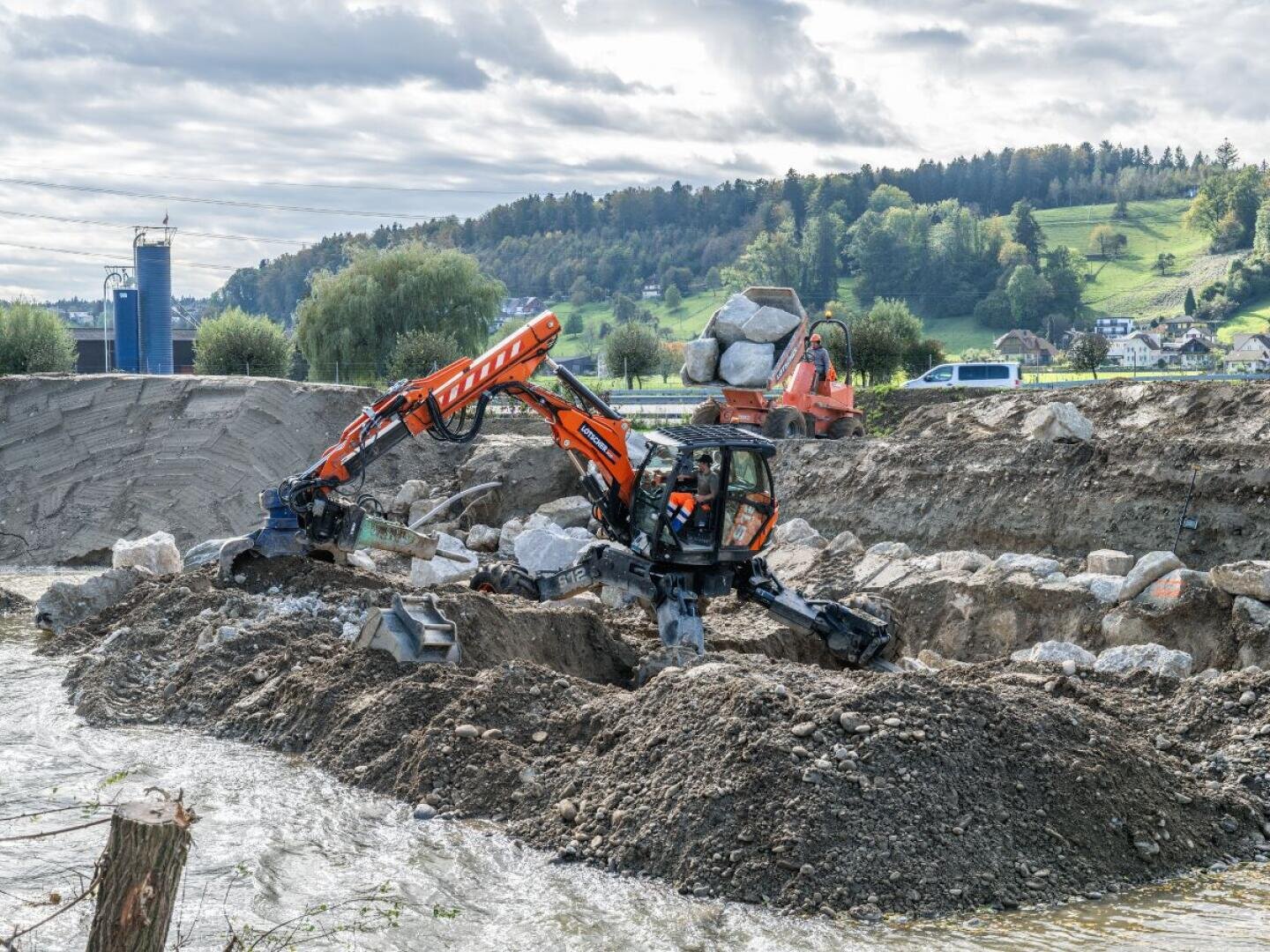 Ein orangefarbener Spinnenbagger bewegt große Steine an einem Flussufer auf einer Baustelle, während ein anderes orangefarbenes Fahrzeug und Steinhaufen in der Nähe zu sehen sind; im Hintergrund sind grasbewachsene Hügel und Bäume zu sehen.
