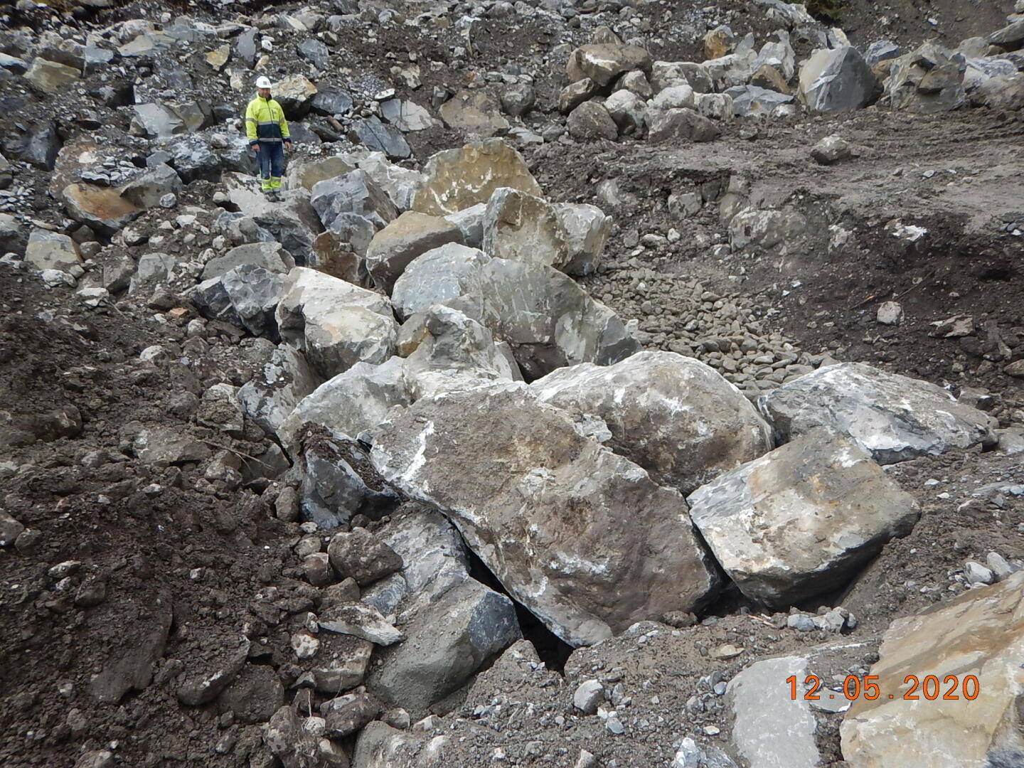 Eine Person mit gelber Sicherheitsjacke und weißem Helm steht zwischen großen, verstreuten Steinen und Felsblöcken in einem zerklüfteten, felsigen Außenbereich. Der Boden ist uneben und es gibt keine Vegetation. Datumsstempel: 12-05-2020.
