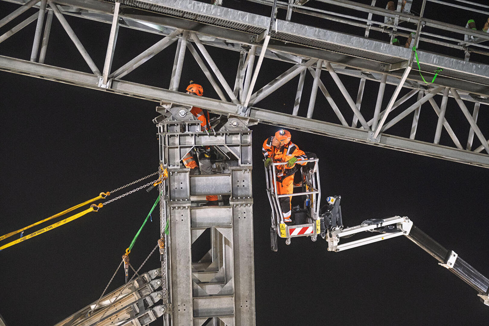 Zwei Bauarbeiter in orangefarbener Schutzkleidung arbeiten nachts an einer Stahlkonstruktion. Der eine steht auf einer Hebebühne, der andere auf einem Gerüst, beide umgeben von Trägern und Sicherungseinrichtungen.