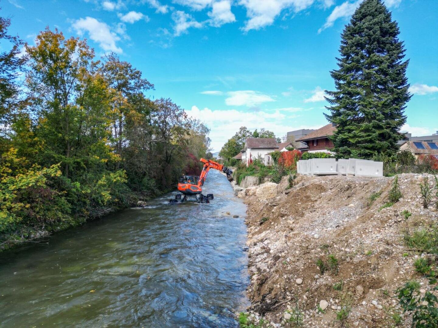 Ein orangefarbener Bagger arbeitet an einem sonnigen Tag in einem schmalen Fluss in der Nähe von Bäumen und Häusern, mit Erdhaufen und Betonblöcken am Flussufer unter einem blauen Himmel mit vereinzelten Wolken.