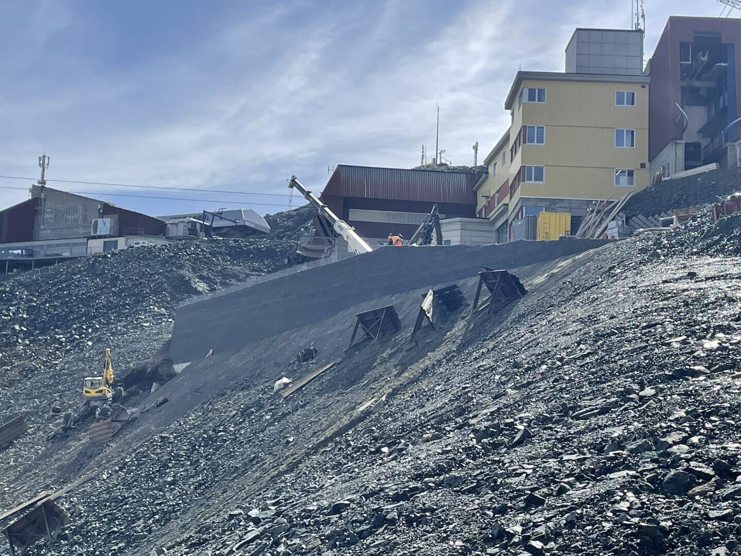 Eine abschüssige, felsige Baustelle mit Maschinen und einem gelben Gebäude im Hintergrund. Der Boden scheint uneben zu sein, und es gibt mehrere in den Hang eingebettete Strukturen. Der Himmel ist teilweise bewölkt.