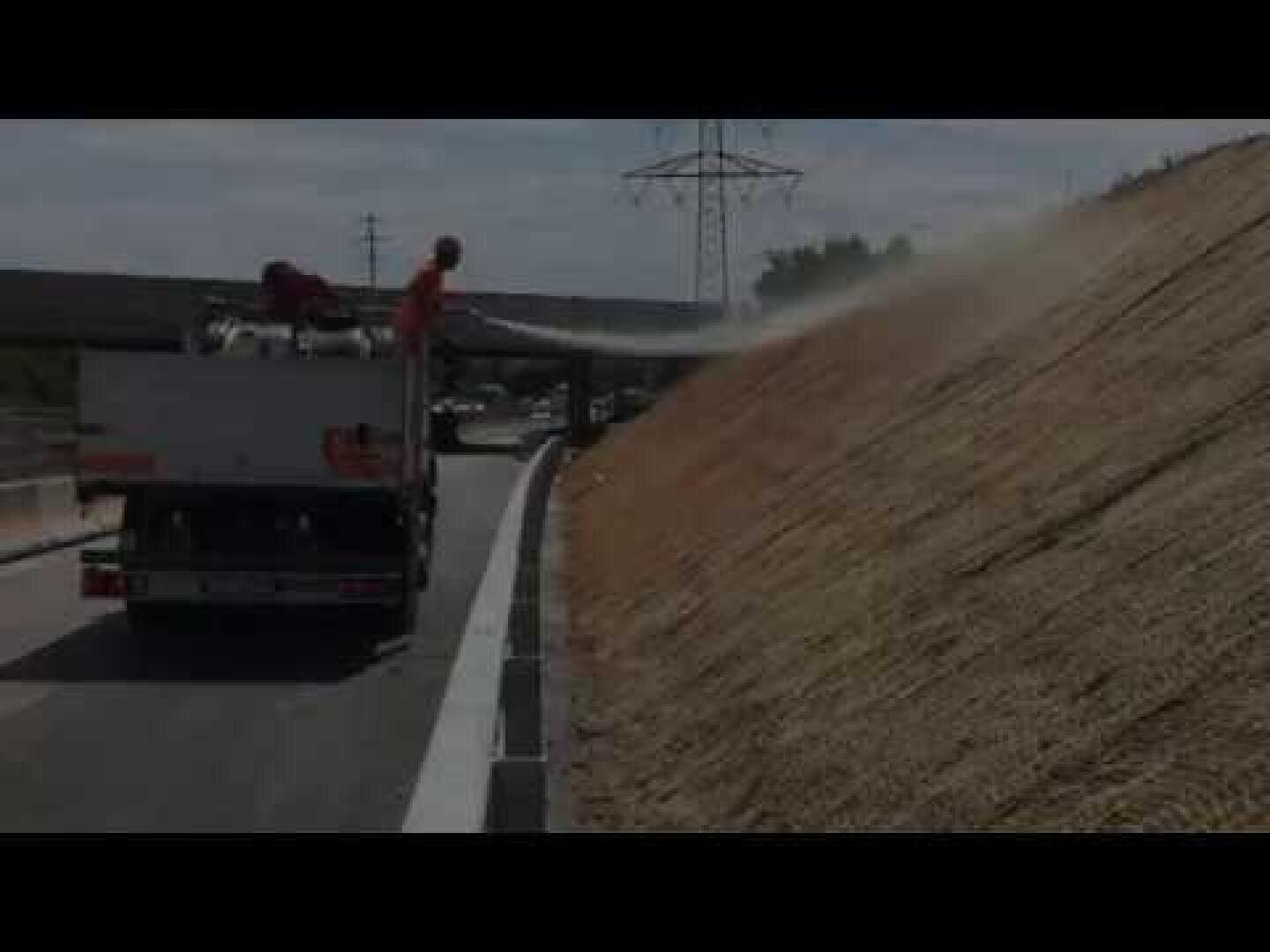 Ein Lkw am Straßenrand sprüht Material auf eine abschüssige Böschung, wahrscheinlich zum Erosionsschutz oder zur Aussaat von Pflanzen; Arbeiter bedienen das Gerät bei teilweise bewölktem Himmel.