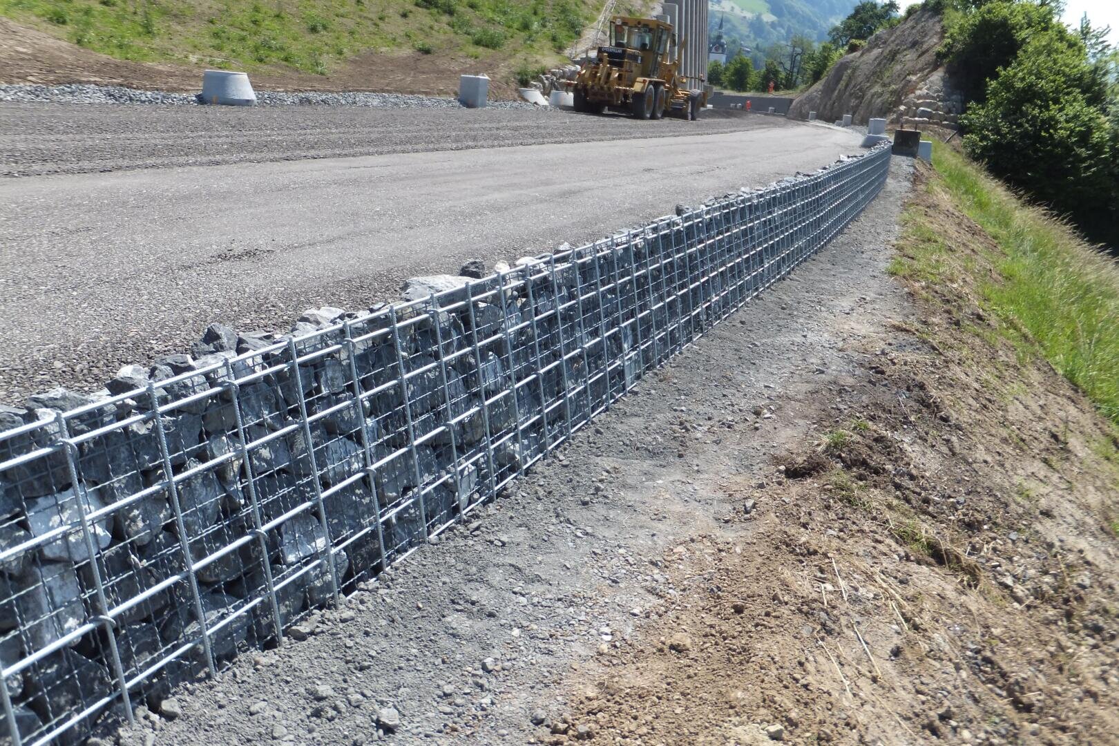 Eine Nahaufnahme einer Gabionenwand, die aus mit Steinen gefüllten Drahtkörben besteht, verläuft entlang einer neu gebauten Straße, während im Hintergrund Baumaschinen zu sehen sind.