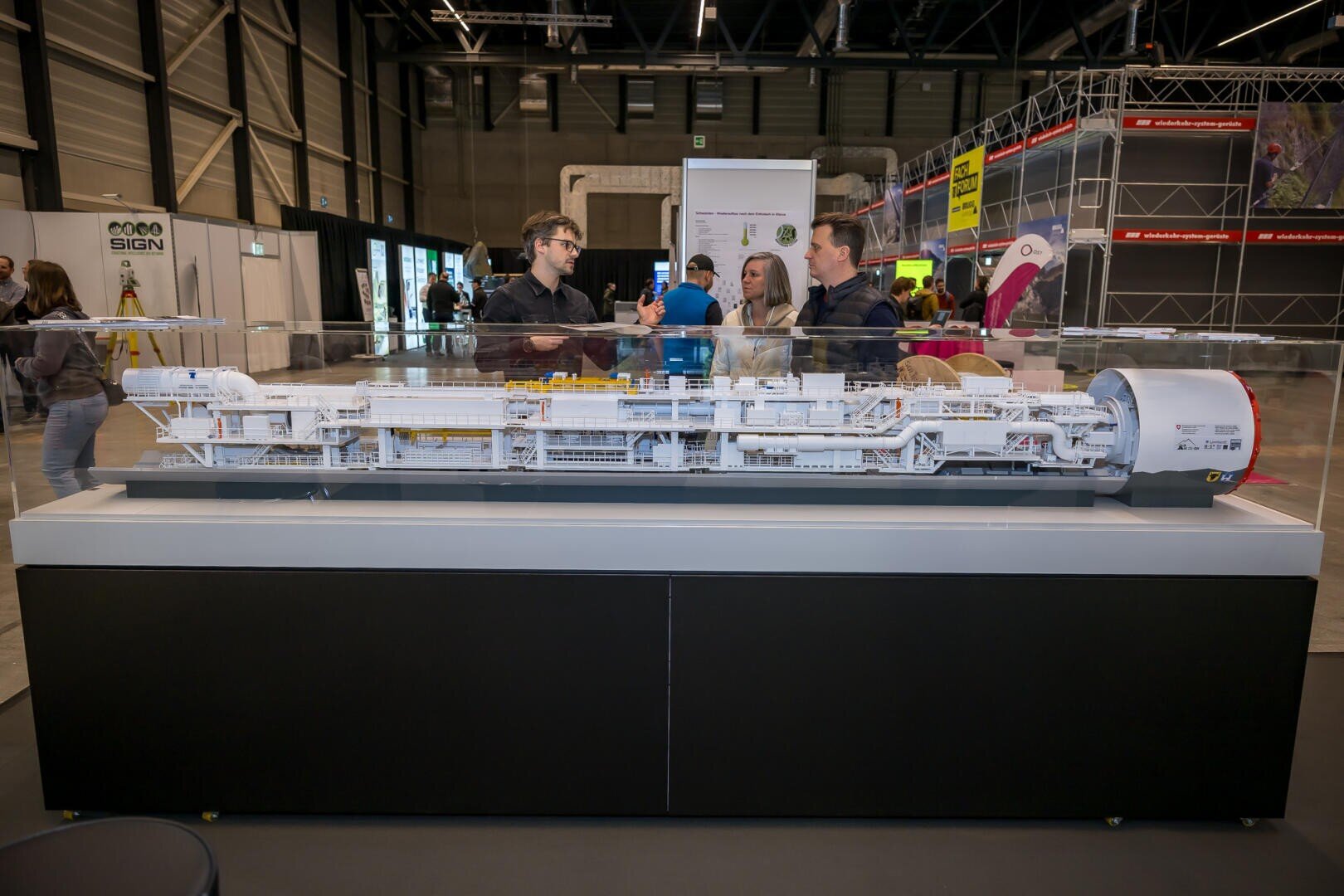 Eine Gruppe von Menschen steht um ein großes, detailgetreues Modell eines Teilchenbeschleunigers, das auf einem schwarz-weißen Tisch in einer Indoor-Ausstellung oder bei einer wissenschaftlichen Veranstaltung ausgestellt ist.