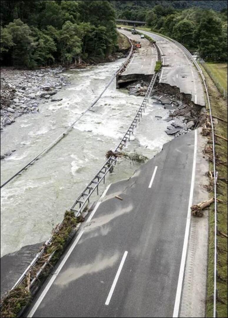 Luftaufnahme eines Flusses, der eine Straße überflutet und schwer beschädigt hat. Große Teile des Gehwegs und der Leitplanken wurden weggespült, Trümmer sind verstreut und Bäume säumen den Schauplatz.