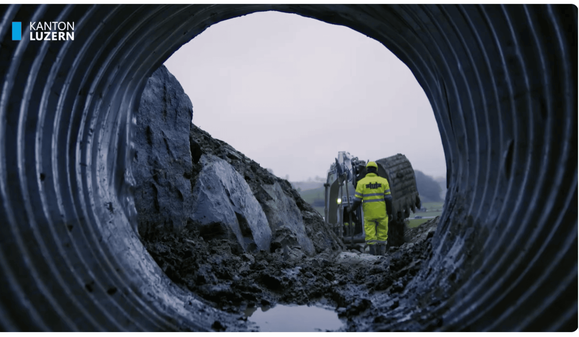 Blick durch ein großes Metallabflussrohr zeigt einen Arbeiter in einem gelben Sicherheitsanzug, der auf schlammigem Boden in der Nähe von Baumaschinen steht; bedeckter Himmel im Hintergrund. Das Logo des Kantons Luzern befindet sich in der oberen linken Ecke.