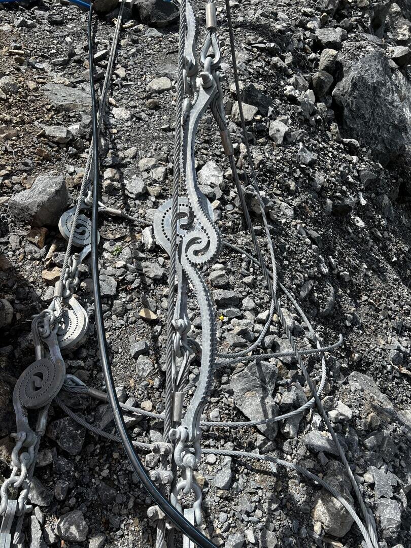 Metallkabelspanner und Drähte sind auf felsigem Boden angeordnet, wobei Zahnräder und Mechanismen sichtbar sind. Die Szene scheint Teil einer industriellen oder baulichen Einrichtung in einer rauen Außenumgebung zu sein.