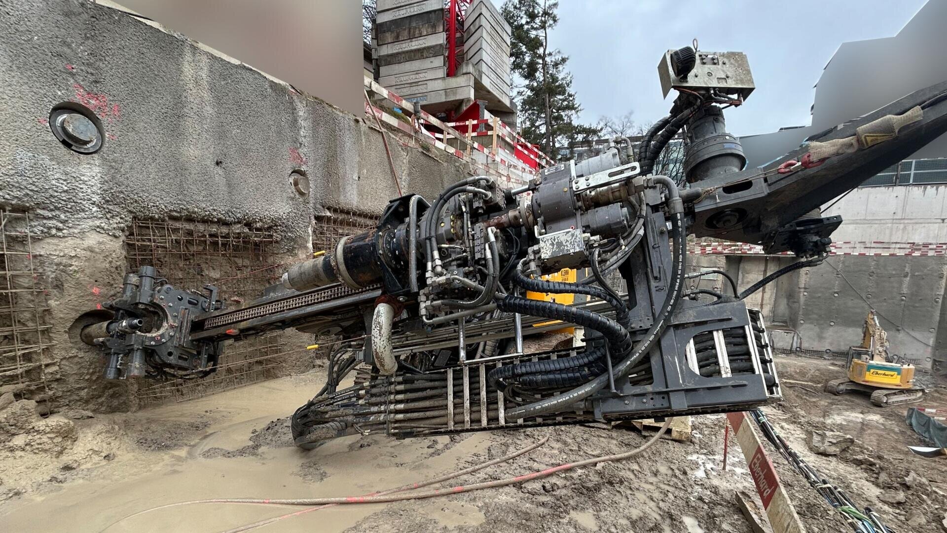 Eine große Bohrmaschine steht waagerecht auf einer Baustelle und bohrt in eine mit Bewehrungsstäben verstärkte Betonwand. Schlamm und Wasser umgeben die Maschine, und im Hintergrund ist Baumaterial zu sehen.